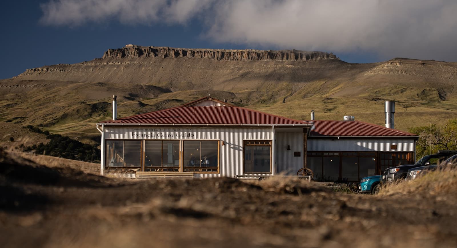 Your base for the week, and the mesa doesn't let you forget where you are. The main lodge building of Estancia Cerro Guido, with white corrugated-iron walls and a red metal roof, set against a broad mesa and green hillside under a partly cloudy Patagonian sky