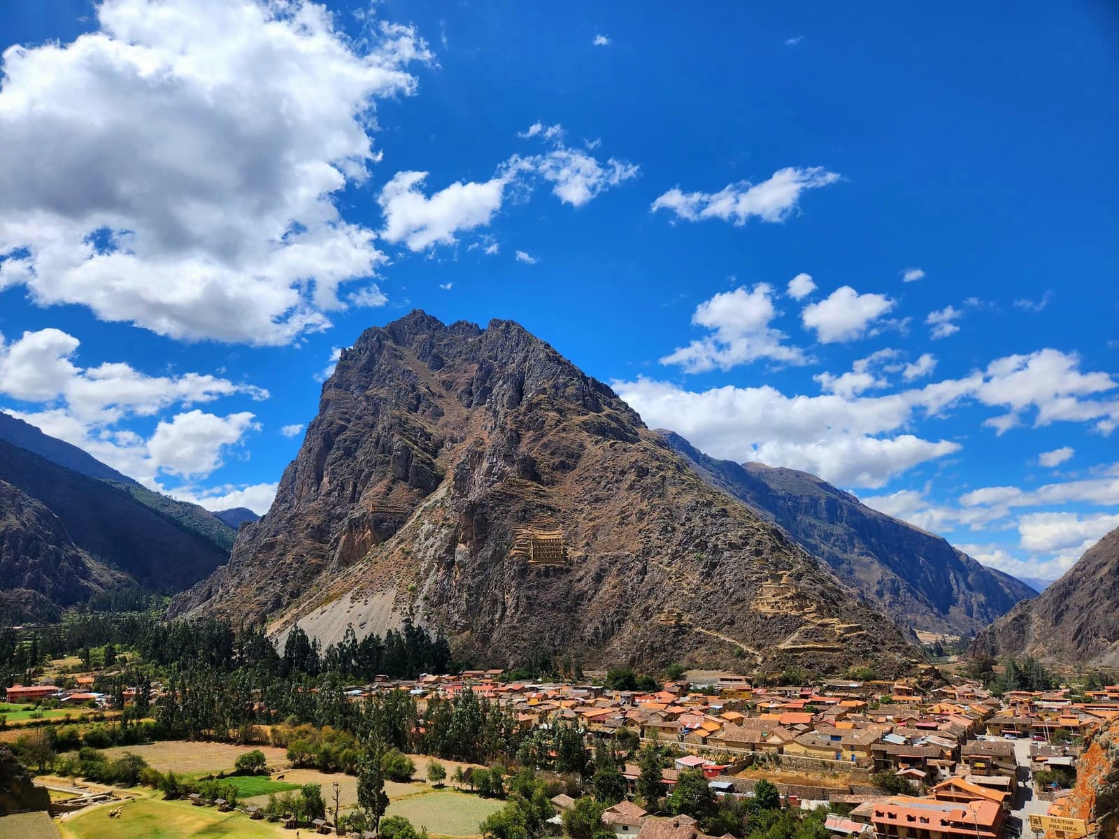 Ollantaytambo from above: the Inca terraces and the modern town share the same mountain, unbothered by the comparison. Panoramic view of Ollantaytambo town with terracotta rooftops spread across the valley floor, ancient Inca terraces carved into the rocky mountain face above, and a bright blue sky with white clouds on this machu picchu amazon tour overland route through Peru