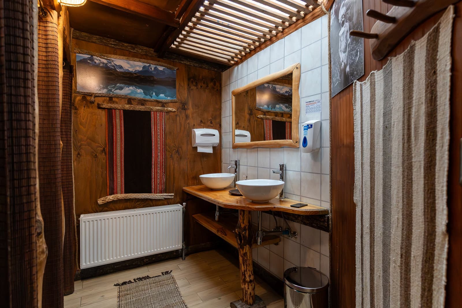 Clean, warm, and a lot more charming than anything you'd expect after nine days on trail. Photo: Standard Dome Shared Bathroom Rustic shared bathroom at EcoCamp Patagonia on the O trek in Patagonia, featuring two white vessel sinks on a live-edge timber vanity, a natural wood-framed mirror, warm wooden walls, and mountain photography prints
