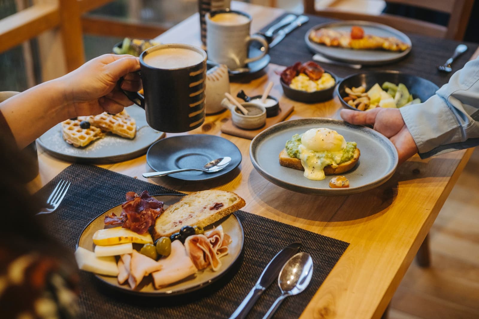 Breakfast here is the kind that makes you genuinely debate skipping the hike. Photo: Patagonia Camp A wooden dining table at Patagonia Camp set with multiple breakfast plates including eggs benedict with hollandaise on avocado toast, a charcuterie and cheese plate, waffles, scrambled eggs, and fresh fruit, with two guests' hands reaching for coffee and a dish