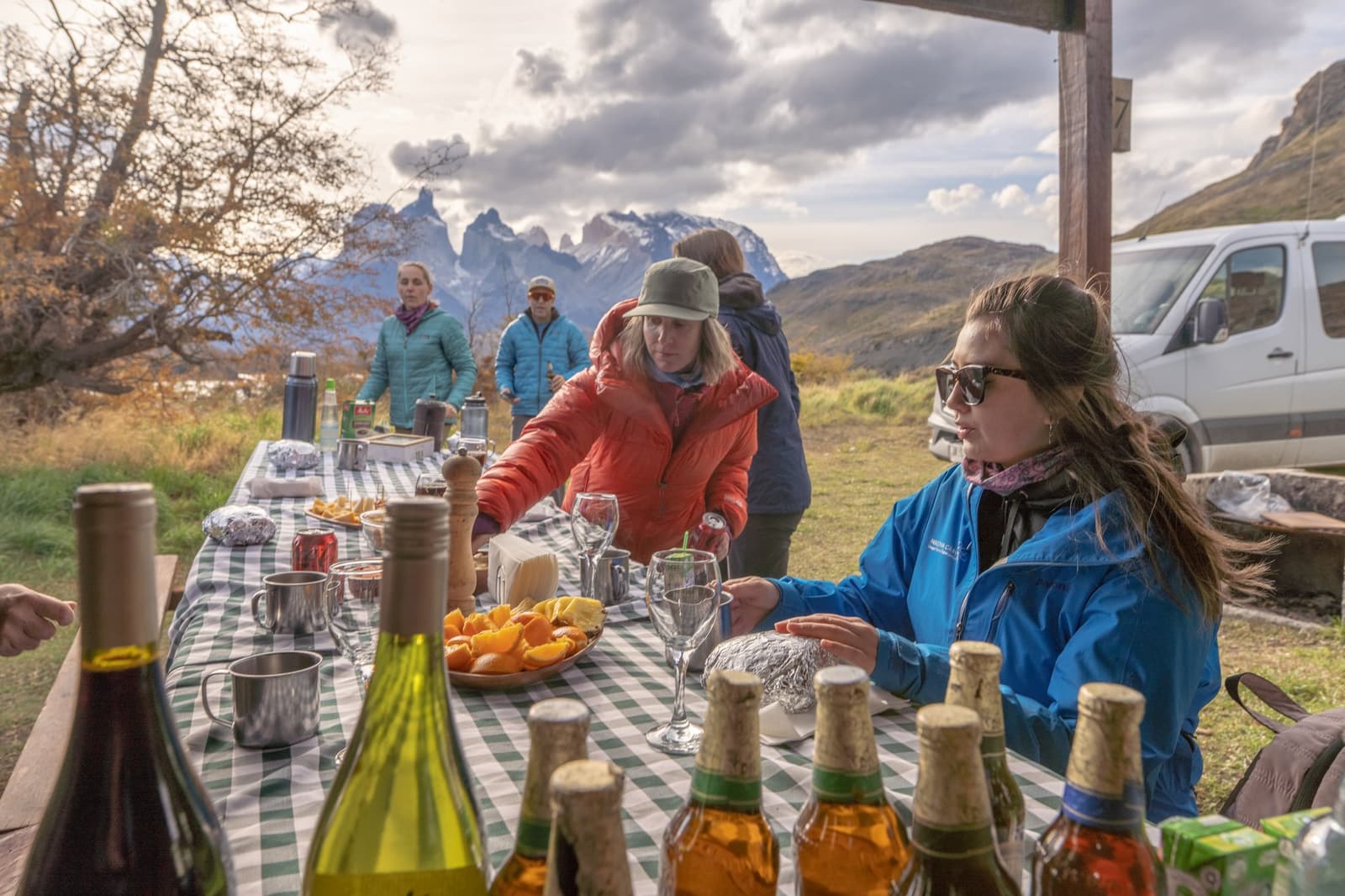 Cold wind, good cheese, cold wine. Some lunches earn their place. Photo: Patagonia Camp A group of travellers gathered around a gingham-cloth picnic table laden with wine bottles, fresh oranges, cheese, and glasses, with the jagged granite peaks of the Paine Massif rising under cloudy skies in the background — a patagonia camp outdoor lunch in the field