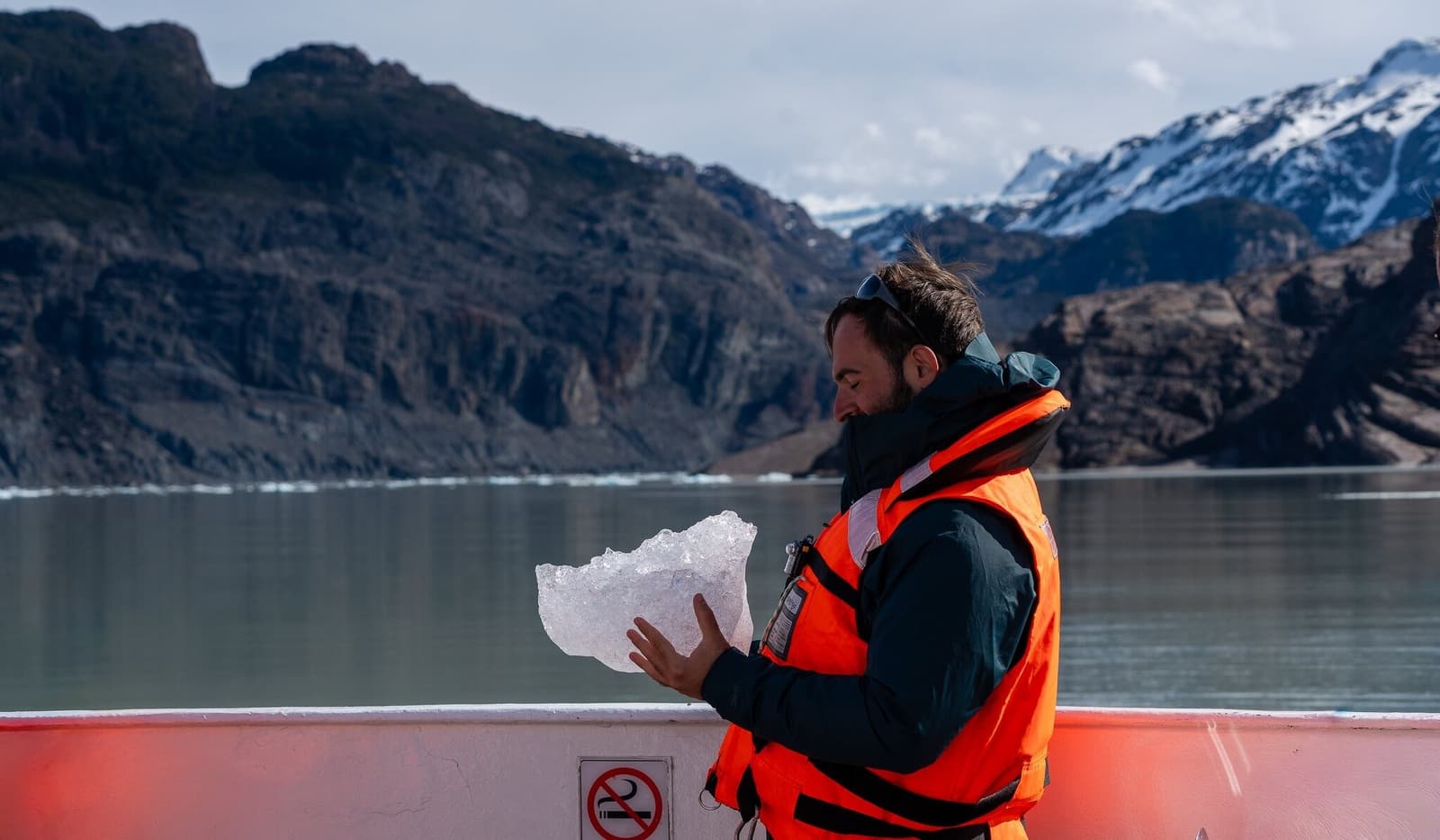 Thousands of years old, cold enough to feel it in your teeth. A bearded traveler in an orange life jacket holds a large chunk of glacial ice on the deck of a boat during the short w trek Grey Glacier excursion, with snow-dusted mountains and a calm grey-green lake visible behind him