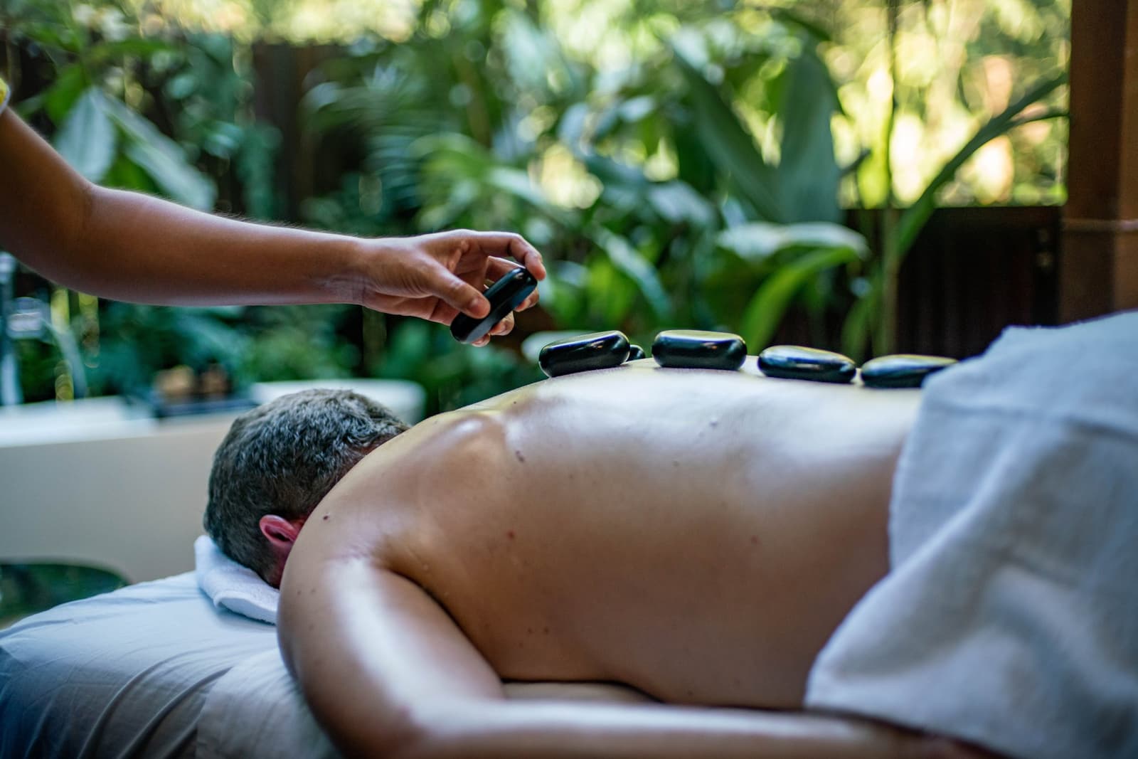 Even deep in the Amazon, your back gets the attention it deserves. A therapist places warm black basalt stones along a guest's back during a hot stone massage at a jungle spa, with dense tropical foliage visible through open walls — a wellness moment at the Tambopata Research Center Peru.