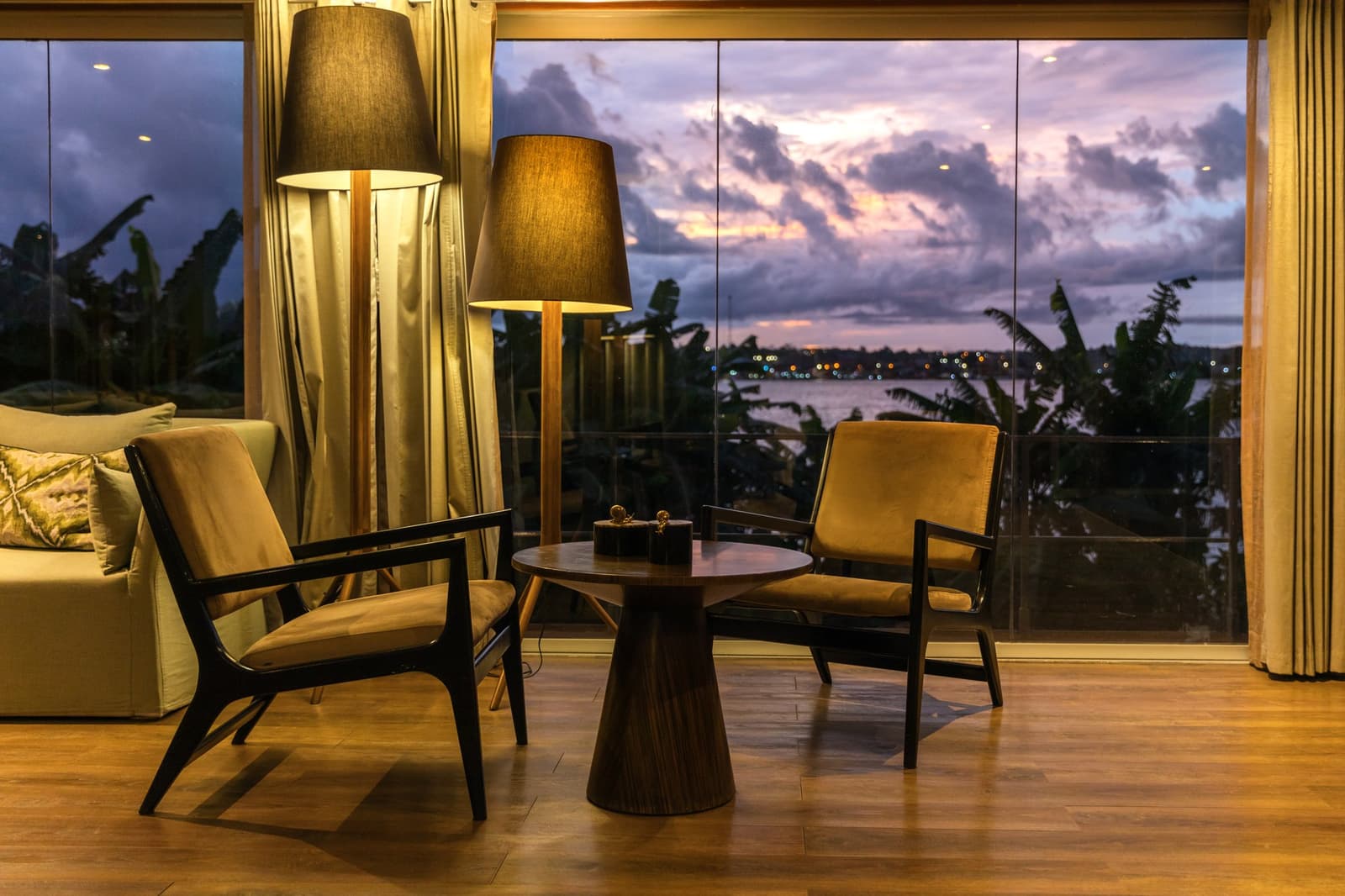 Two chairs, one river, and a sky that does all the talking. Photo: Yvonne Kreuzmayr Warmly lit lounge seating area on the Aria Amazon cruise, with two mid-century armchairs and a round wooden table set against floor-to-ceiling panoramic windows overlooking a river and a dramatic purple and orange dusk sky