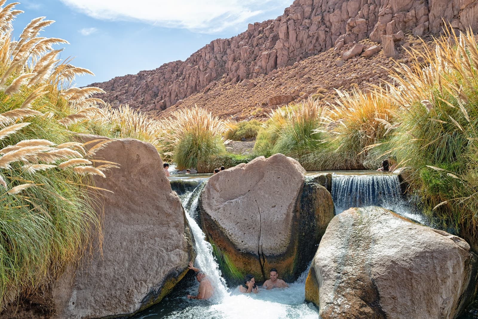 Hot water tumbling over boulders, cold desert air above. Hard to leave. Bathers soaking between large boulders in the warm cascading pools of Puritama Hot Springs, surrounded by golden pampas grass and red rock canyon walls, a highlight of this atacama desert tour package