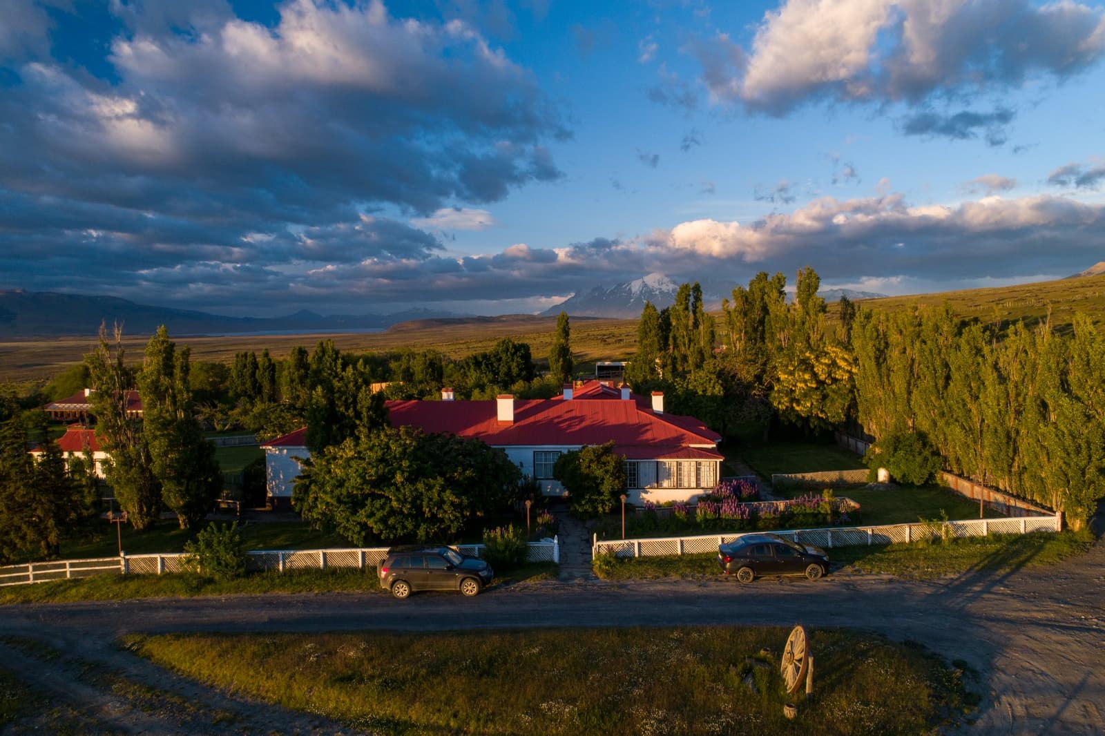 Poplars, lupines, a red roof, and a mountain that keeps changing its mind about the weather. Aerial view of Estancia Cerro Guido at golden hour, showing the white manor house with a red roof surrounded by tall poplar trees, lupine gardens, and open Patagonian steppe stretching toward a snow-capped peak under a dramatic blue sky with large clouds