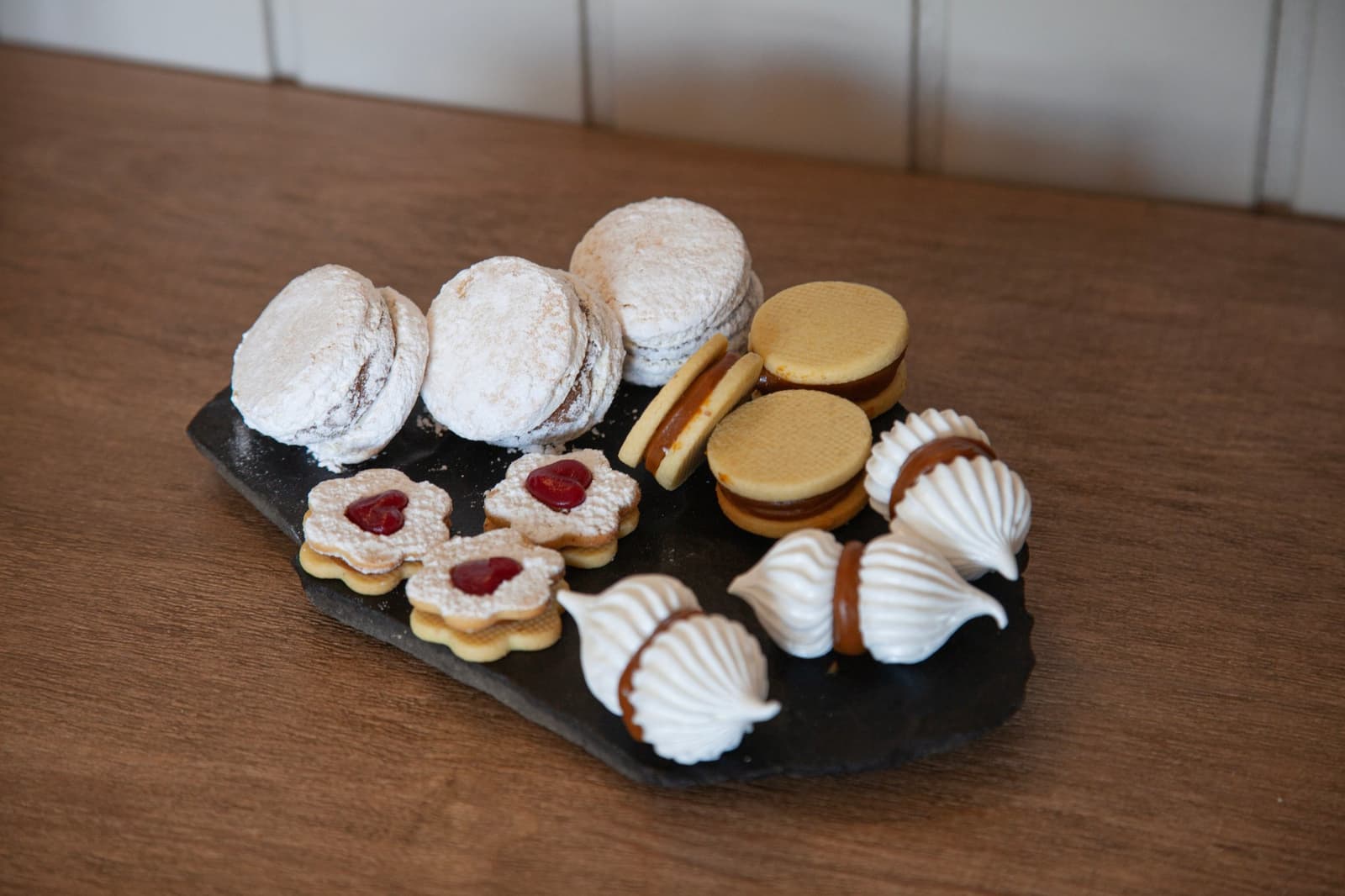 Four kinds of cookies, one slate board, zero complaints. A slate board at Estancia Cerro Guido holds an assortment of house-made cookies including powdered-sugar sandwich biscuits, round wafer alfajores with caramel filling, flower-shaped linzer cookies with red jam centers, and white meringue shells drizzled with dulce de leche