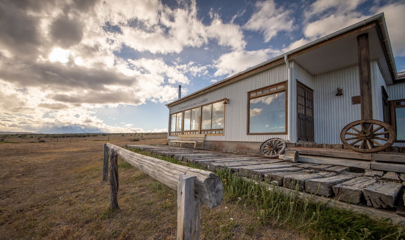 A century of wind, wool, and good company written into every plank. The white corrugated-iron facade of Estancia Cerro Guido, a patagonian estancia operating since 1906, with a weathered wooden boardwalk, vintage wagon wheels leaning against the wall, and the open steppe stretching toward distant mountains under a dramatic clouded sky