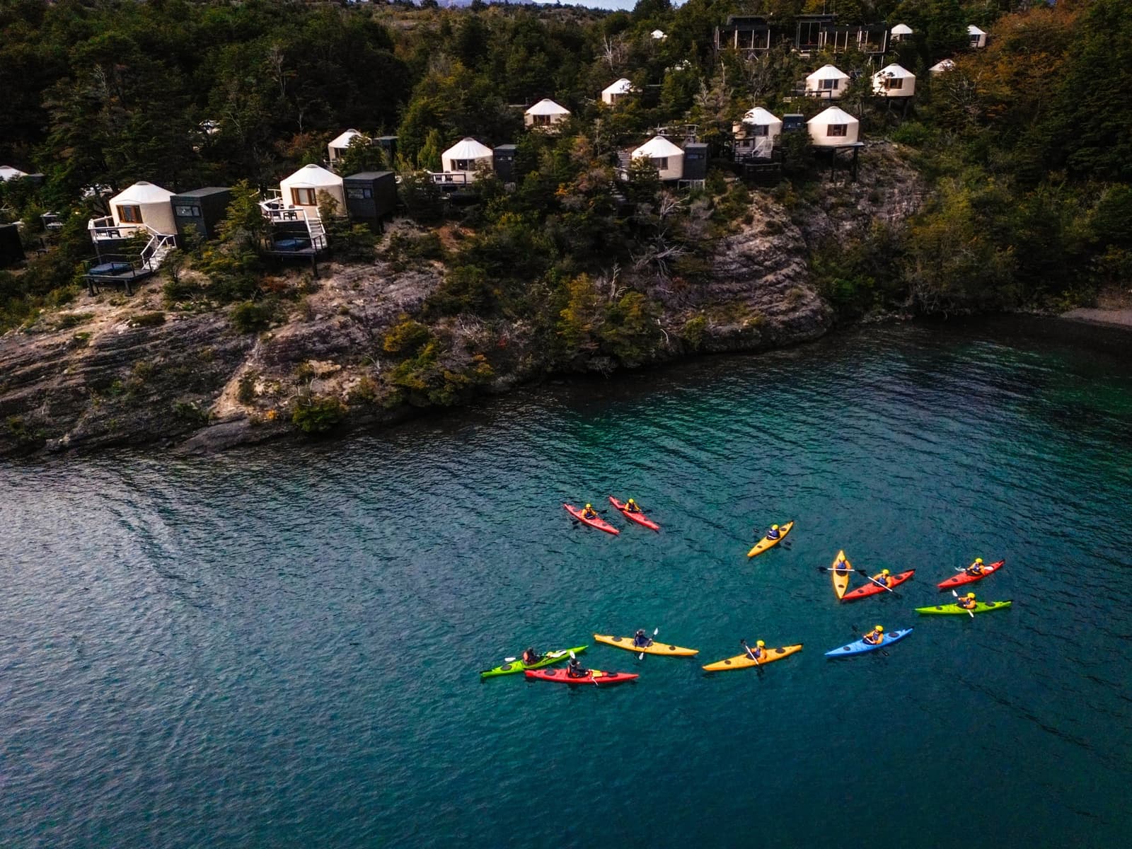 Eleven kayaks, one lake, zero wrong turns, the yurts waiting right above. Photo: Patagonia Camp Aerial view of a group of paddlers in colorful kayaks on the turquoise waters of Lake Toro, with Patagonia Camp's white luxury yurts nestled among native forest on the rocky shoreline above
