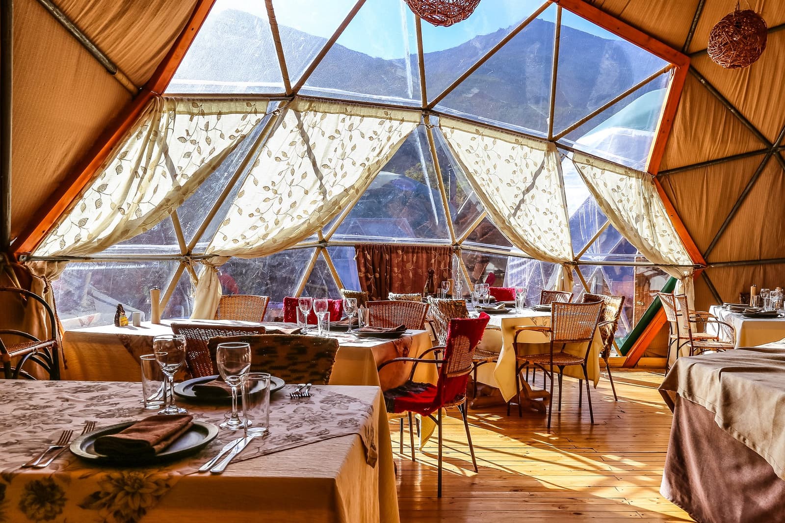 Jantar com vista para a montanha, sem precisar reservar. Photo: Community Dome Interior de um restaurante em cúpula geodésica na Torres del Paine W Trek, com mesas postas, cadeiras de vime, cortinas bordadas e uma crista de montanha visível através dos painéis triangulares transparentes