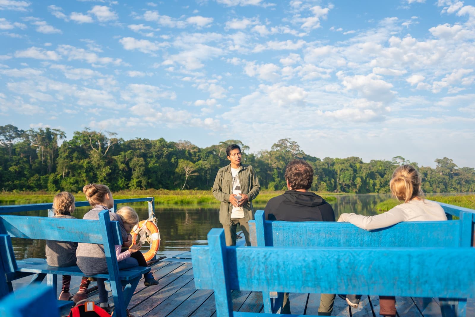 Good guides make the forest talk. This one clearly has a lot to say. A local naturalist guide addresses a small group of travellers seated on blue wooden benches aboard a river boat, with dense Amazon rainforest and calm water visible in the background, during an amazon tour from puerto maldonado