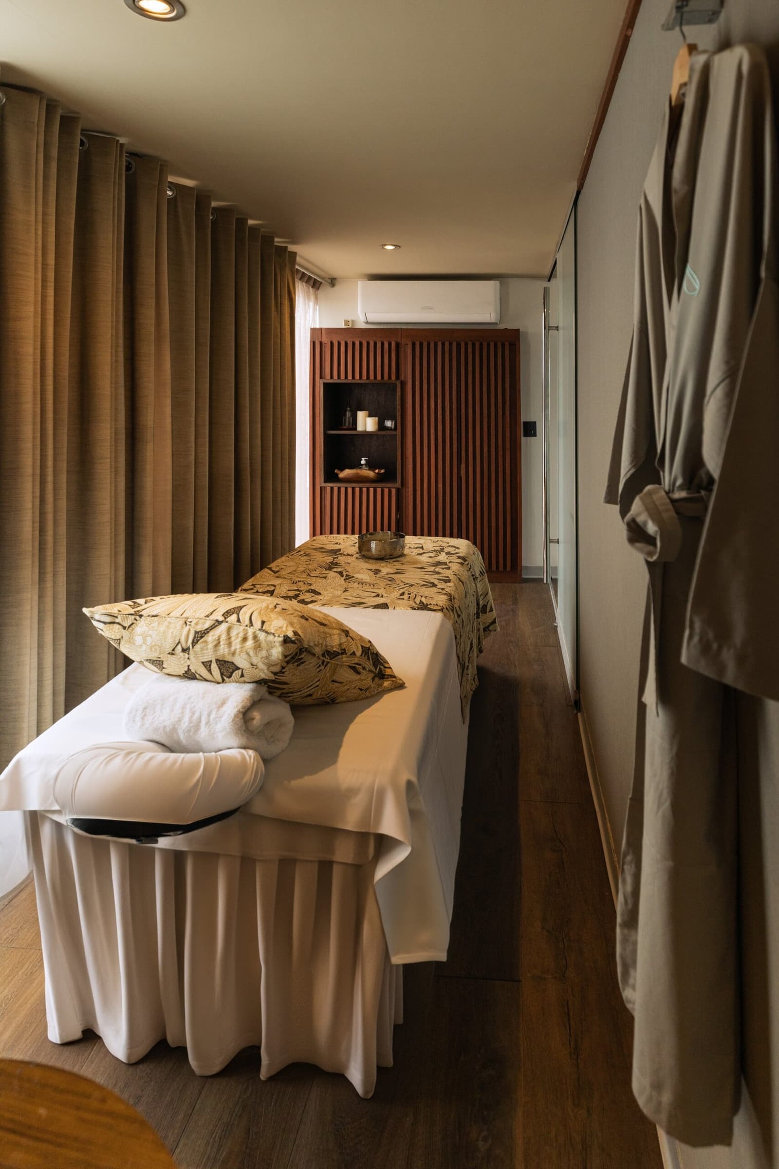 The jungle can wait. This room has other plans for you. Photo: Yvonne Kreuzmayr A prepared massage treatment room on the Aria Amazon cruise, with a white-draped table dressed in botanical-print pillows, folded towels, a small bowl, warm wood slatted cabinetry, linen curtains, and a spa robe hanging on the wall