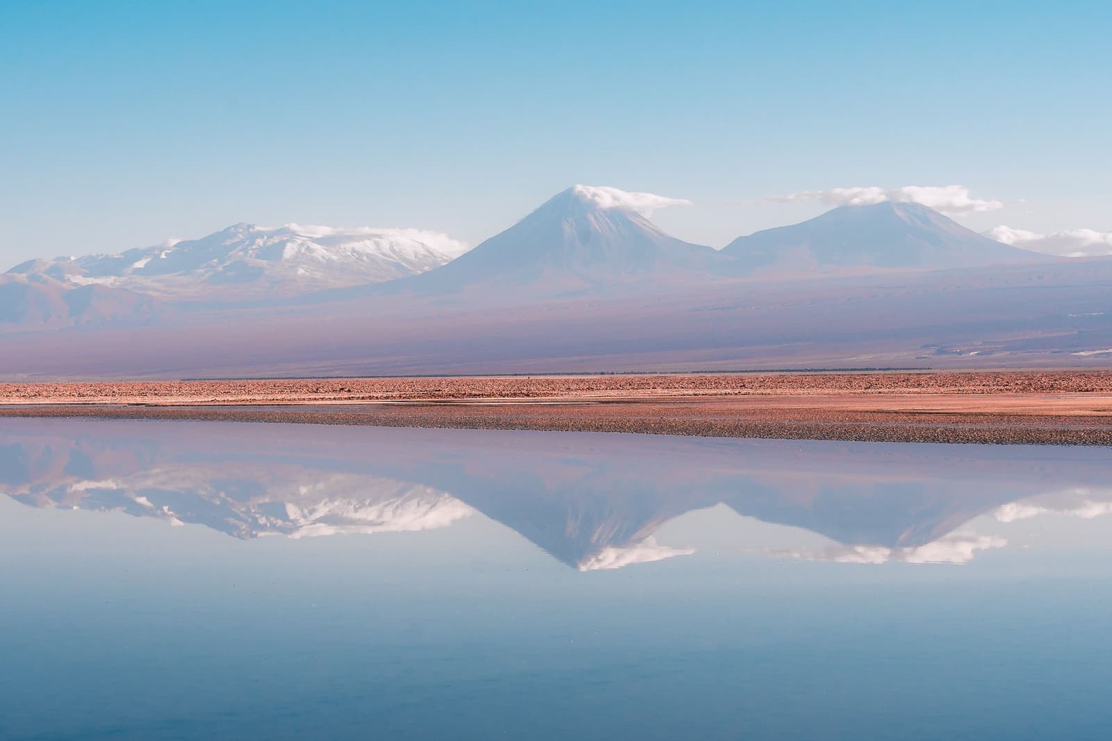 Still water, and suddenly the desert has two skies. Snow-capped Andean volcanoes perfectly mirrored in the glassy surface of a shallow salt flat on this atacama desert tour package, with rust-colored desert flats stretching across the middle ground under a clear blue sky