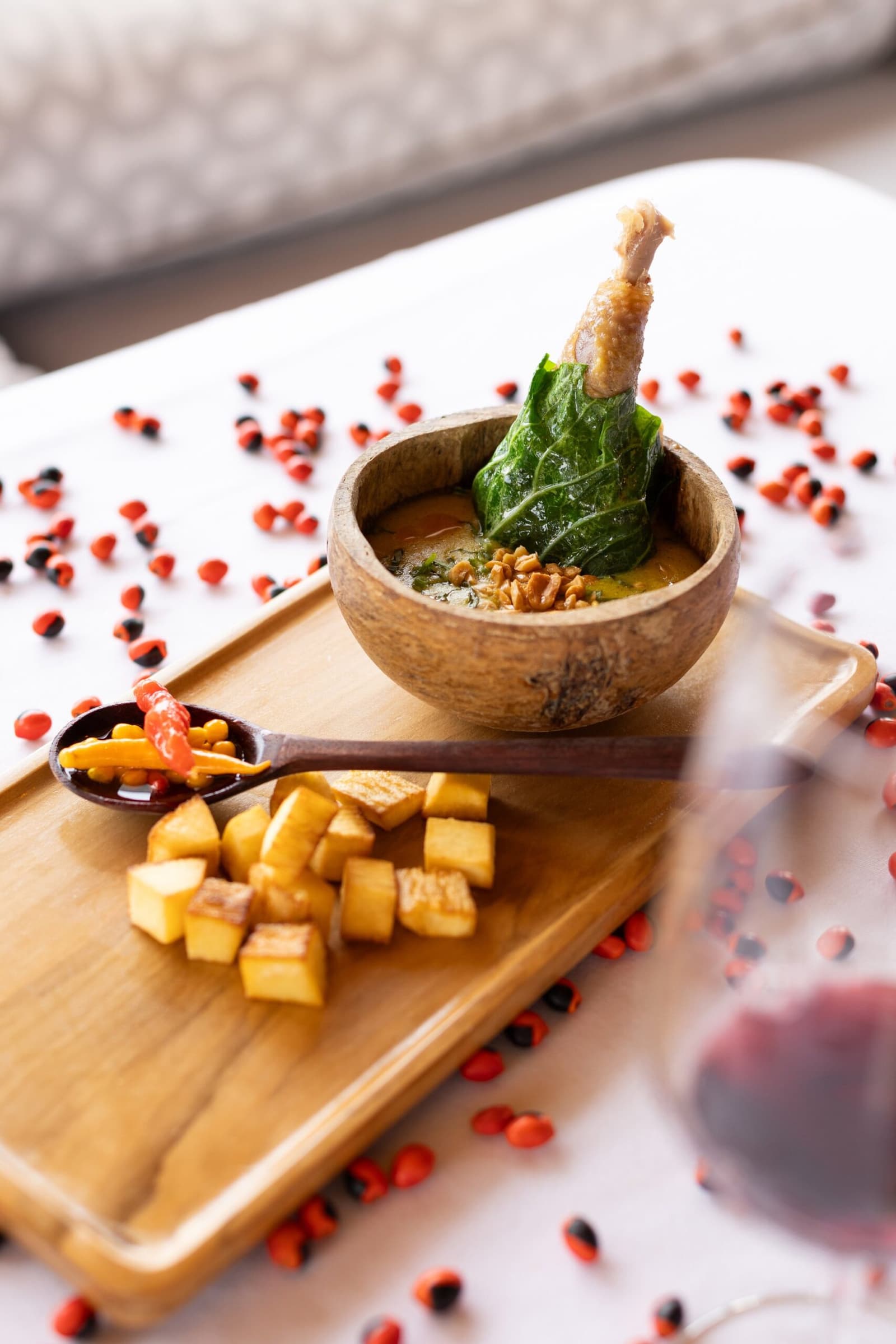 Cozinha regional confiante o suficiente para espalhar sementes pela mesa assim, com estilo. Uma tigela de madeira com caldo dourado, coxa de ave braseada, folhas murchas e grãos tostados servida em uma bandeja de madeira com cubos de legumes assados e uma colher escura com pimenta e milho, parte da gastronomia do cruzeiro Delfin Amazon