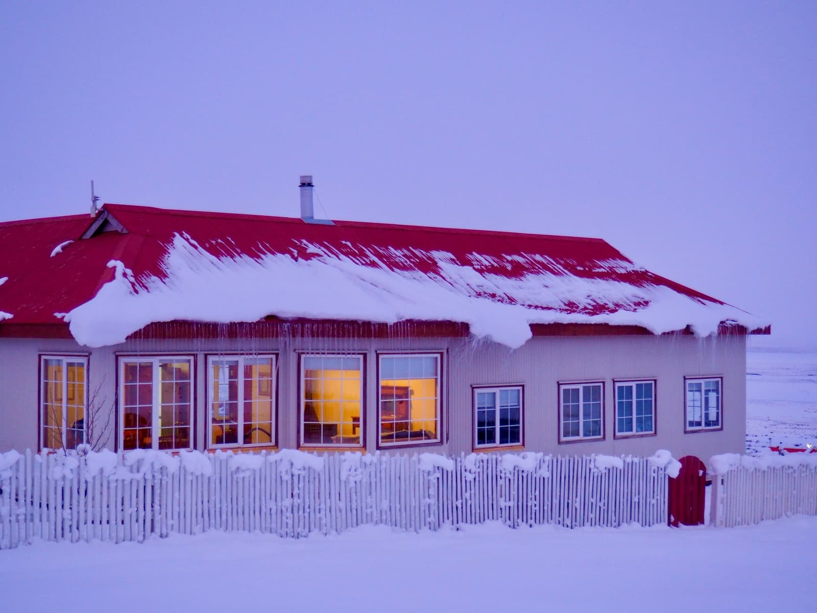 Even in deep winter, the lights inside are on and the welcome is real. Estancia Cerro Guido's main building at dusk in winter, with a snow-laden red metal roof, icicles hanging from the eaves, a snow-covered white picket fence, and warm amber light glowing through large windows against a pale violet sky
