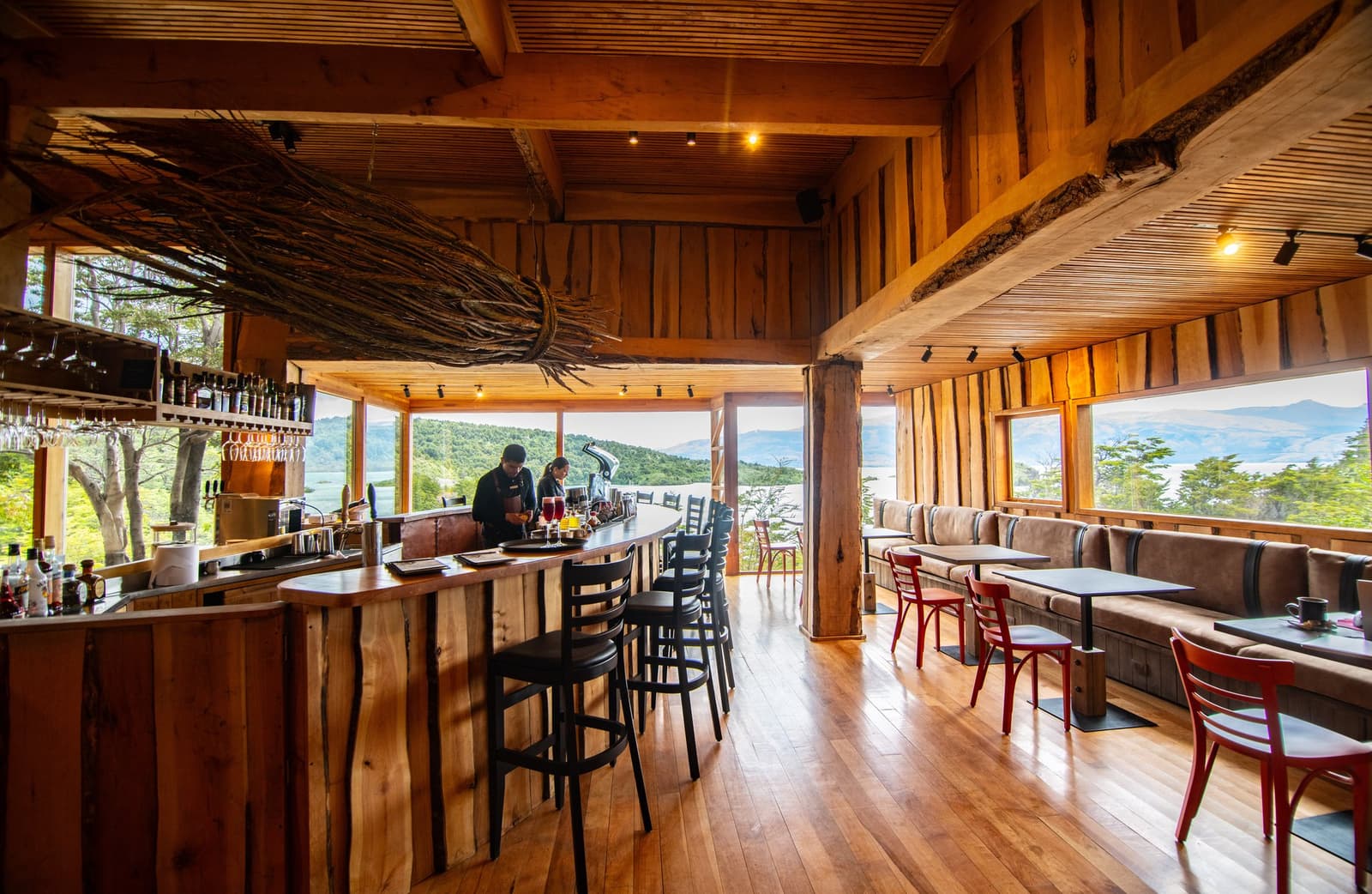 Bar open, lake in the window, and someone already knows what you need. Photo: Patagonia Camp Patagonia Camp's timber-clad bar and lounge, where two staff prepare drinks at a live-edge wooden counter beneath a sculptural driftwood ceiling installation, with panoramic windows framing Lake Toro and the surrounding green hills beyond