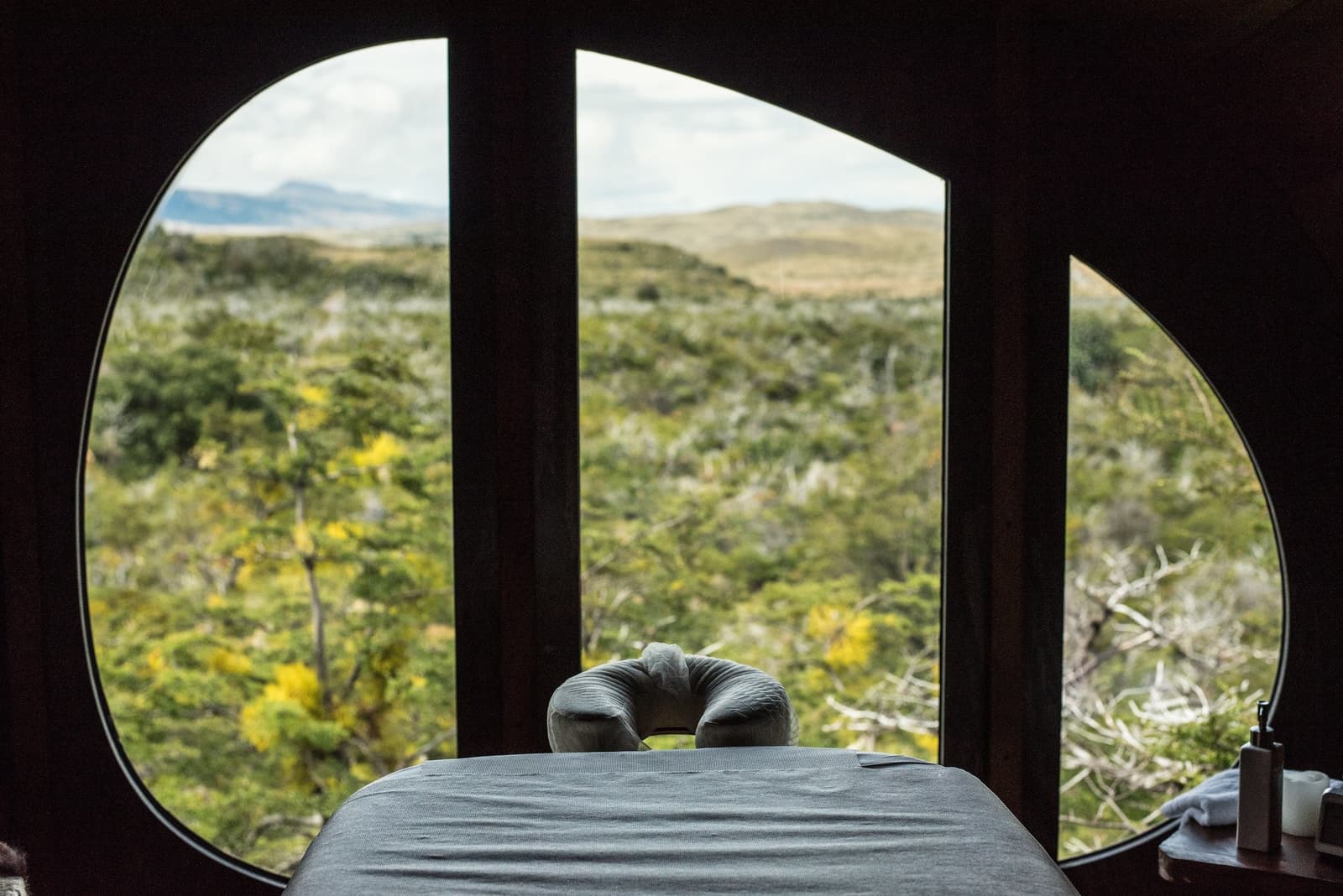 A good massage with a Patagonian wilderness view, because you earned it. A massage table with a face cradle sits inside an eco-lodge spa room, framing a large organic-shaped window that looks out over dense native scrubland and distant hills on a torres del paine 4 day all inclusive tour