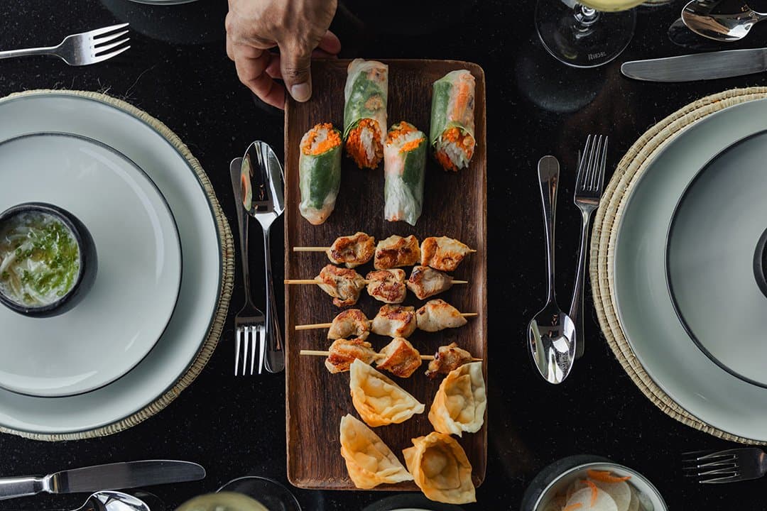 Three courses on one board, zero hesitation about where to start. Photo: Yvonne Kreuzmayr Overhead view of a wooden sharing board on the Aria Amazon cruise featuring fresh spring rolls, grilled meat skewers, and golden fried dumplings, flanked by two elegantly set place settings on a dark table