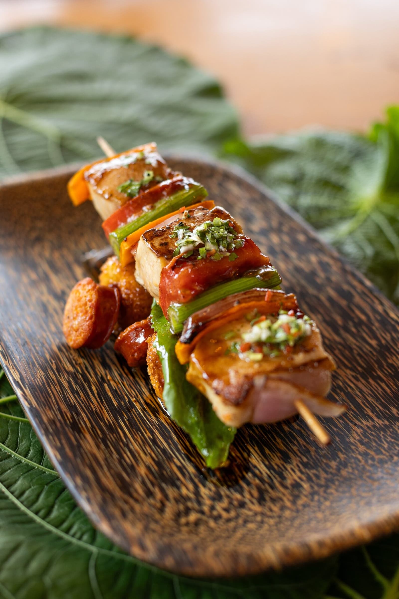 Defumado, dourado na brasa e finalizado com ervas. O almoço aqui não é coisa simples. Um espeto grelhado de carne, pimentões e legumes cobertos com gremolata de ervas, servido em um prato de madeira escura com folhas tropicais