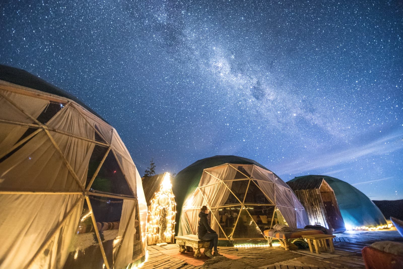 Luz quente, céu frio, e a galáxia fazendo sua parte bem acima do seu domo. Photo: Community Dome Três domos geodésicos iluminados do EcoCamp Patagônia na W Trek de Torres del Paine, com uma figura solitária sentada entre eles sob a Via Láctea no céu noturno