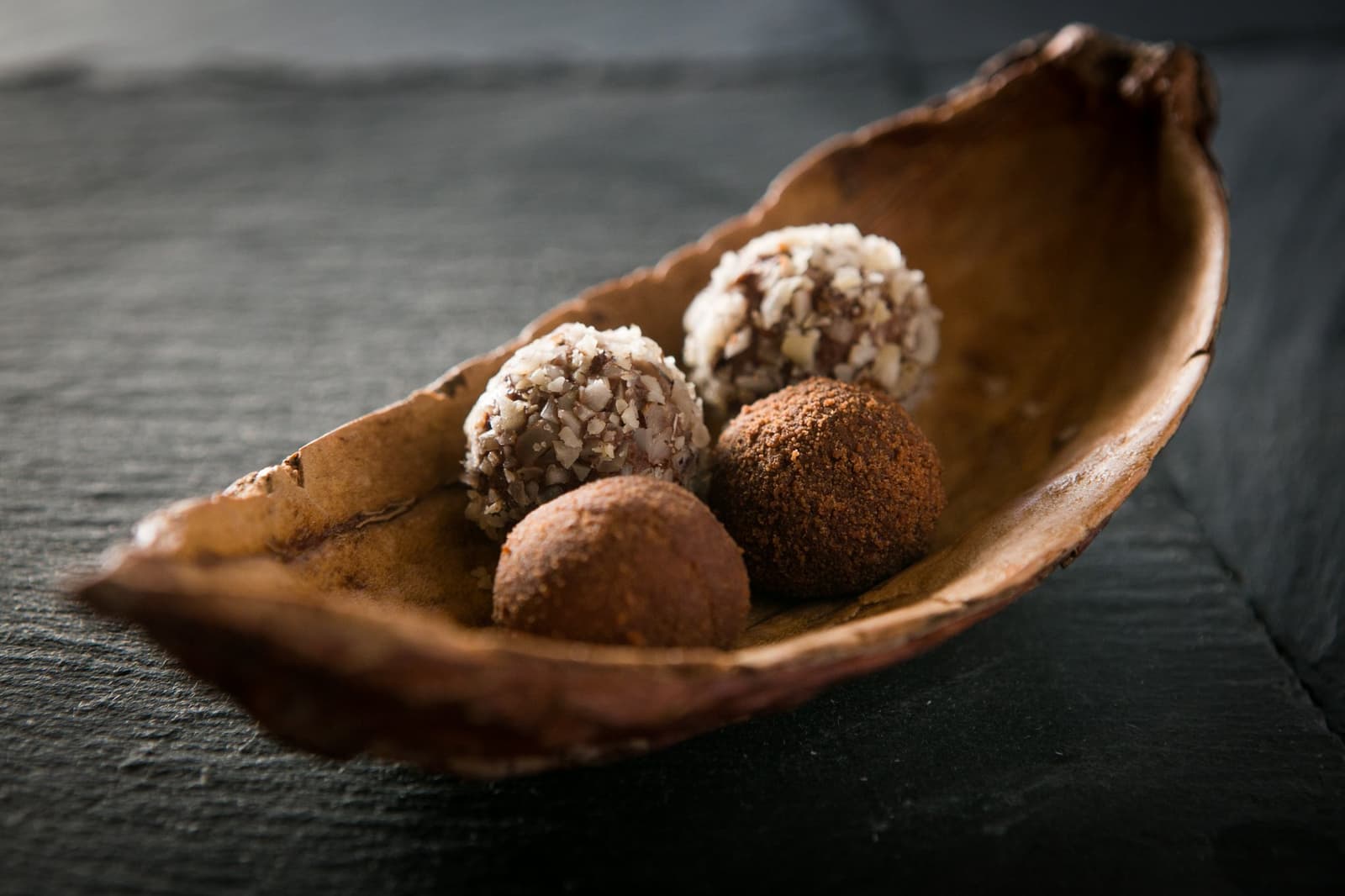 Petit fours that taste like the jungle had a say in the recipe. Four handcrafted chocolate truffles — two rolled in shredded coconut and two dusted with cocoa powder — served in a rustic dried-pod bowl on a dark slate surface, part of the all-inclusive dining experience on the Aria Amazon cruise