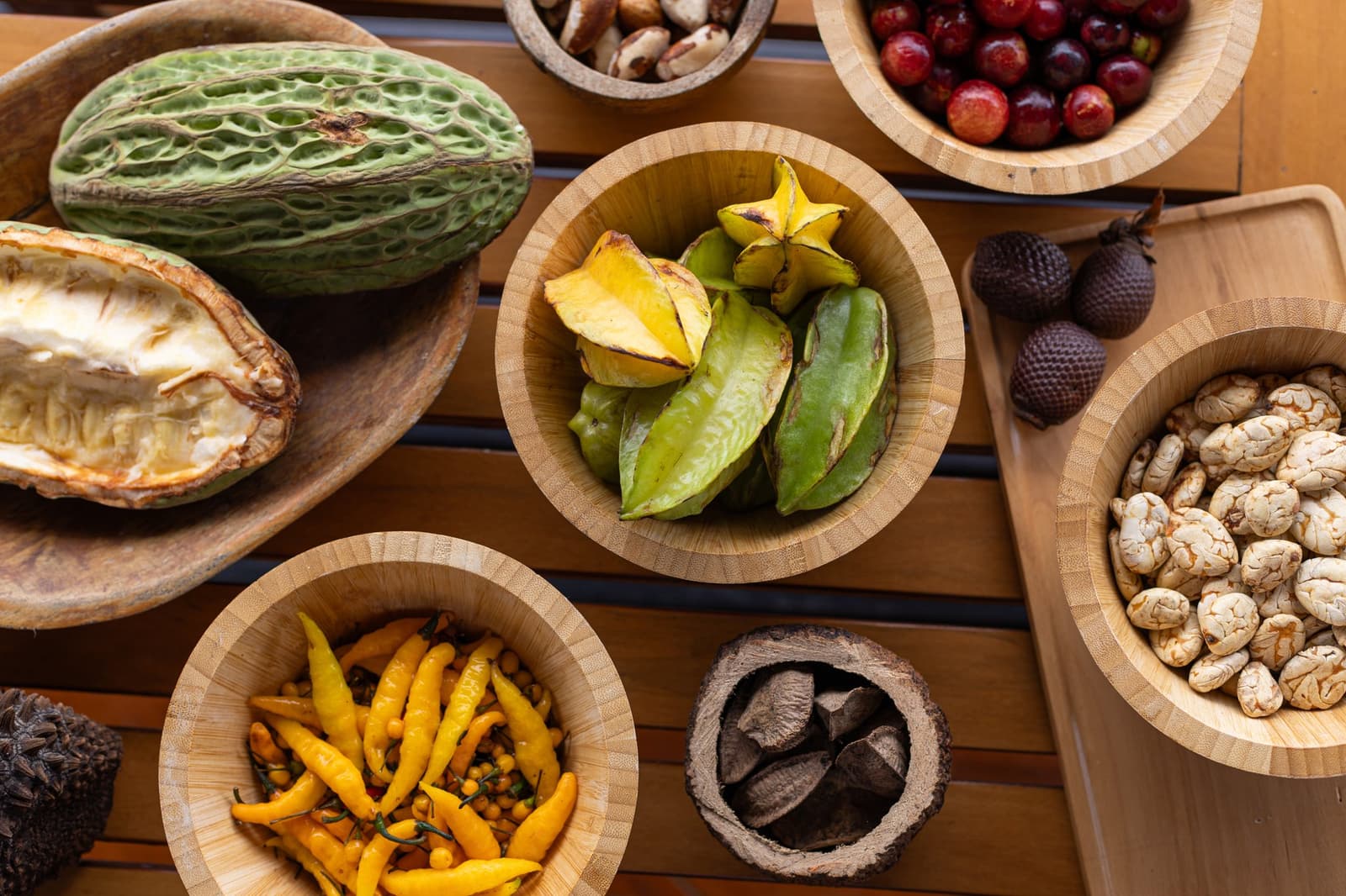 Uma aula de biodiversidade servida à mesa, sem precisar de livro. Vista aérea de frutas, nozes e sementes amazônicas em tigelas de madeira sobre uma mesa ripada, incluindo um cacau partido ao meio, carambola, pimentas amarelas, castanhas-do-pará, pequenas frutas vermelhas e amêndoas de cacau secas