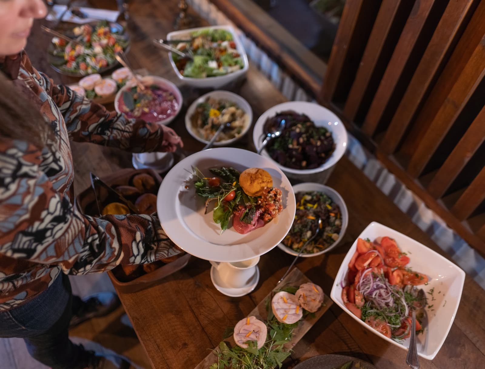 A table this full is its own kind of welcome to Patagonia. Overhead view of a wooden dining table at Estancia Cerro Guido laden with colorful salads, grain dishes, cherry tomato salad, garnished appetizers on a cake stand, and a guest in a patterned shirt reaching to serve