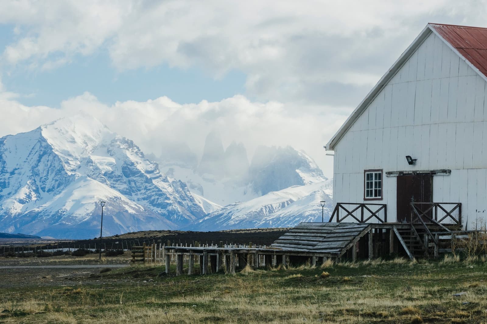 Old wool country, new purpose, and those peaks keeping watch in the background. Photo: Tomas Encina Retamal A white timber farmhouse with a red roof and weathered wooden loading platform stands in open grassland at estancia cerro guido, with snow-covered peaks of the Paine Massif rising behind it under a cloudy sky