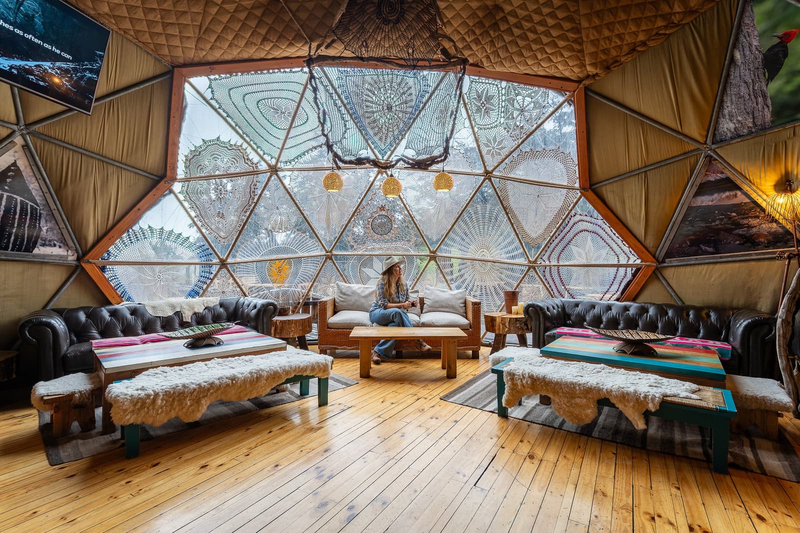 Crochet lace on geodesic glass: EcoCamp's common dome has its own sense of style. Photo: Community Dome Interior of a geodesic dome lounge at EcoCamp Patagonia, with large triangular windows decorated with crocheted lace panels, leather chesterfield sofas, sheepskin-covered low tables, and a woman in a wide-brim hat seated on a wicker sofa — the base for the o trek patagonia o trek in patagonia