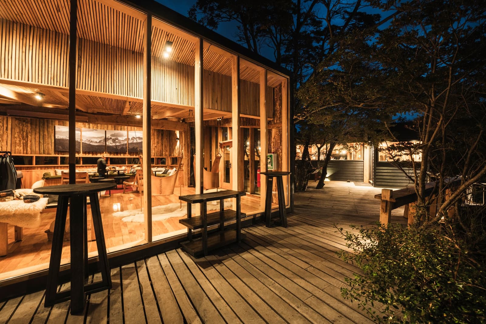 Glass walls, warm wood, and the whole evening ahead of you. Photo: Patagonia Camp Patagonia Camp's glass-walled common room glows with warm light at dusk, its wooden slatted ceiling and hanging chair visible from the timber deck outside, surrounded by native trees under a deep blue sky