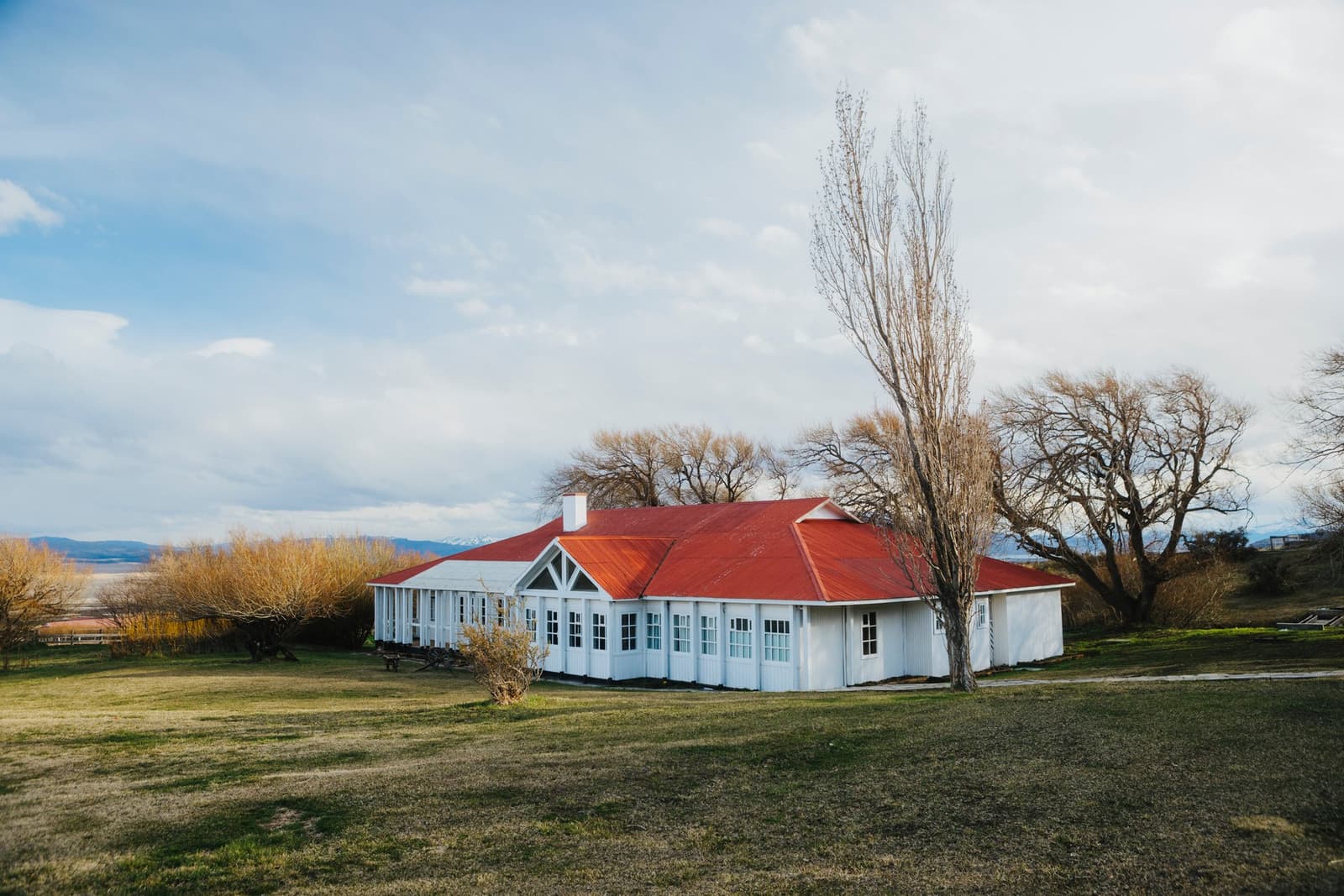 Built to last a century of Patagonian wind, and still doing exactly that. Photo: Tomas Encina Retamal A white-painted ranch building with a red corrugated iron roof at Estancia Cerro Guido, surrounded by bare poplar and willow trees on an open green lawn, with snow-dusted mountains visible in the distance under a partly cloudy sky.