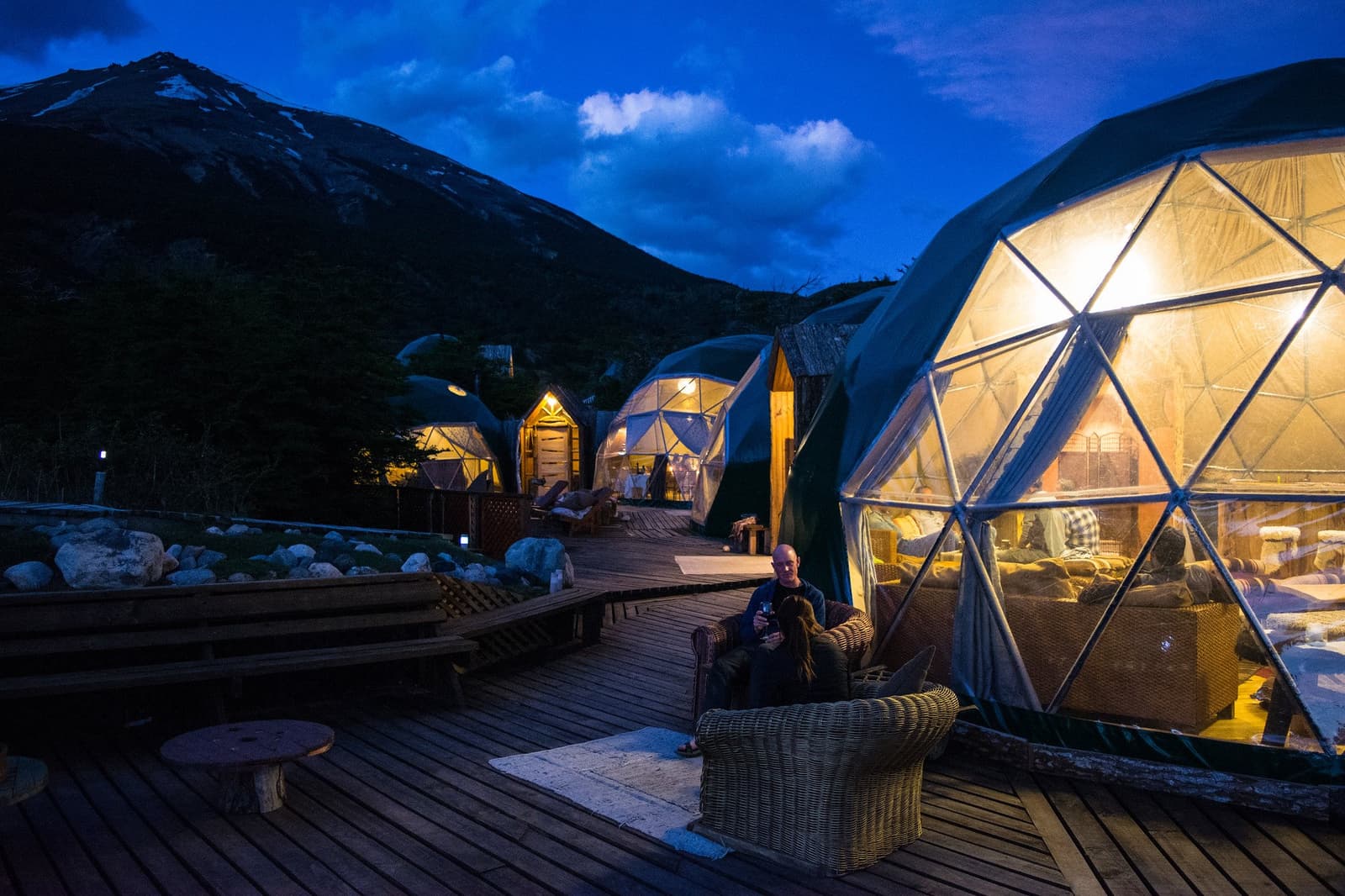 A drink on the deck, the domes lit warm, the mountain going dark. Good evening. Photo: Community Dome A guest relaxes in a wicker chair on the wooden deck of EcoCamp Patagonia at dusk, surrounded by glowing geodesic domes set against a dark mountain silhouette during the O Trek in Patagonia