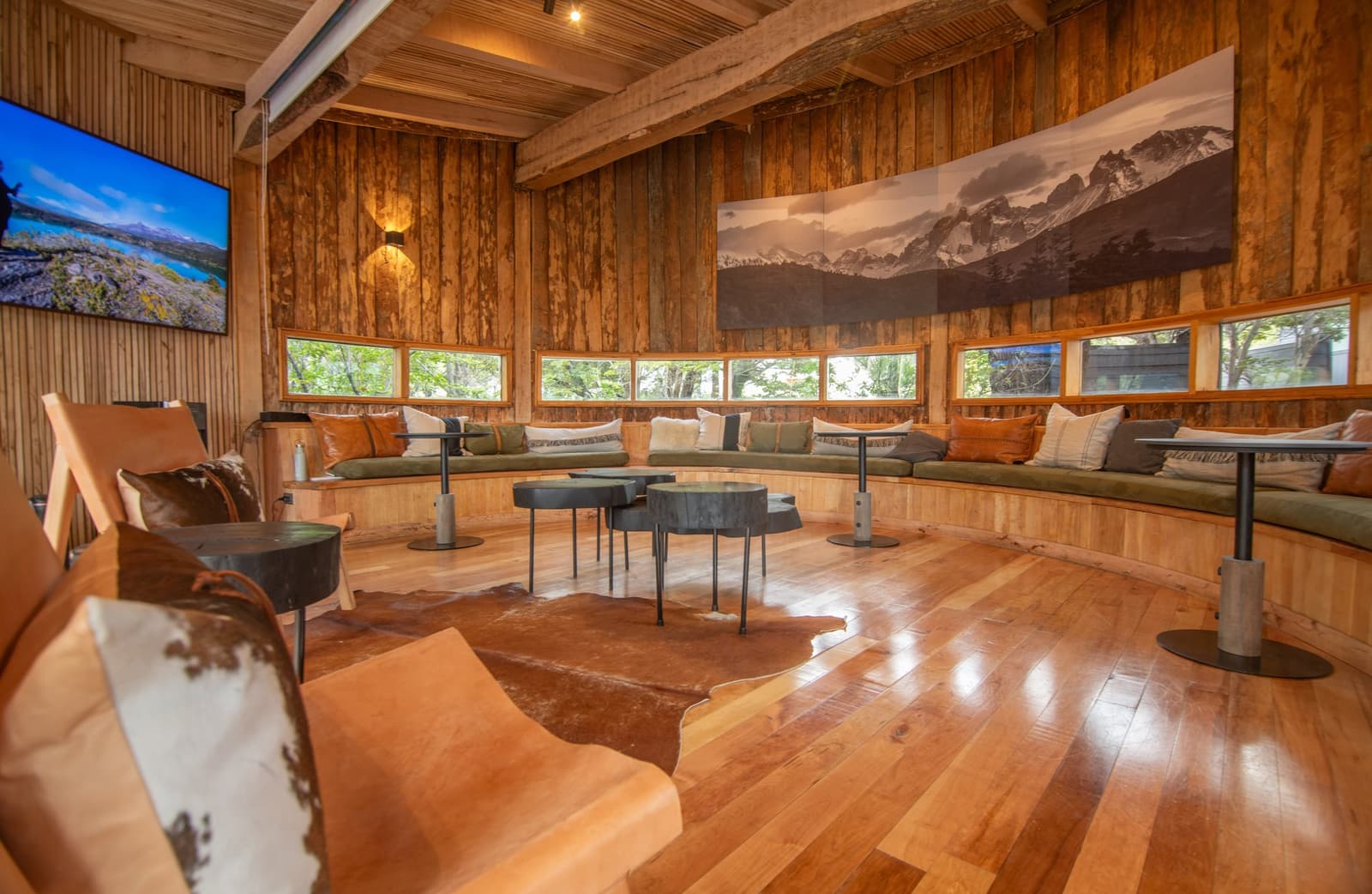 Settle in here after a long day on the trail, no further argument needed. Photo: Patagonia Camp Patagonia Camp's curved communal lounge with polished hardwood floors, a wraparound built-in sofa with green cushions and leather throw chairs, a cowhide rug, and a large panoramic mountain photograph mounted on wood-panelled walls