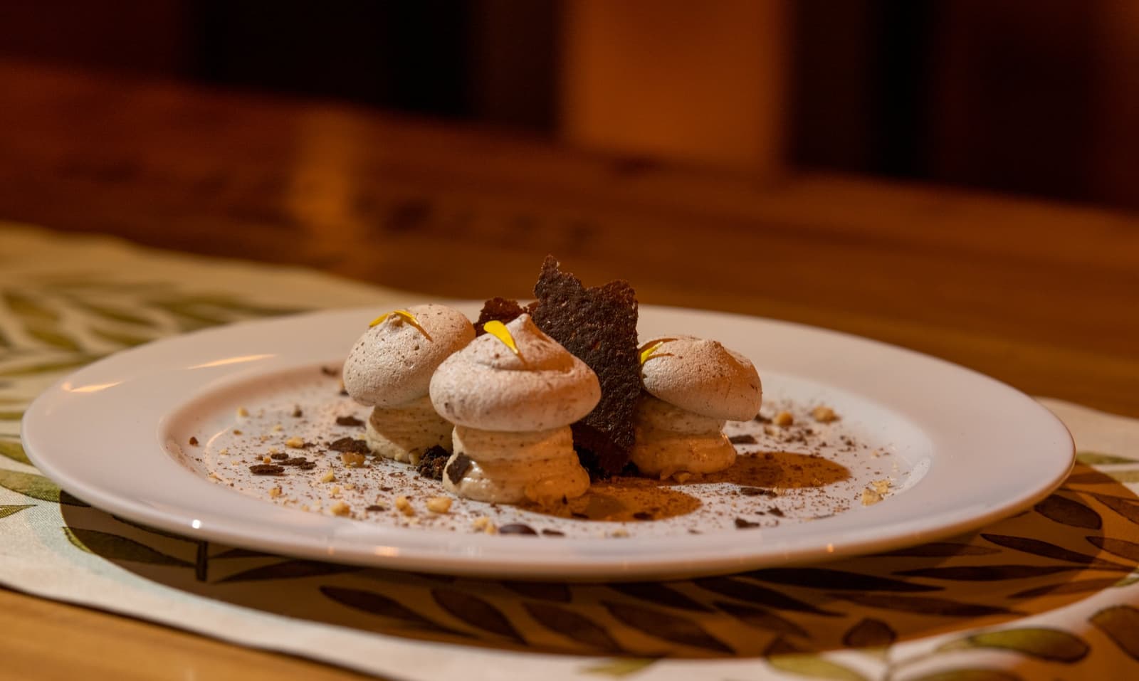 Meringue, chocolate, petals. The kitchen out here has range. Plated dessert at Estancia Cerro Guido featuring three cocoa-dusted meringue kisses, a dark chocolate tuile, crushed nuts, and yellow flower petals on a white plate set on a wood table