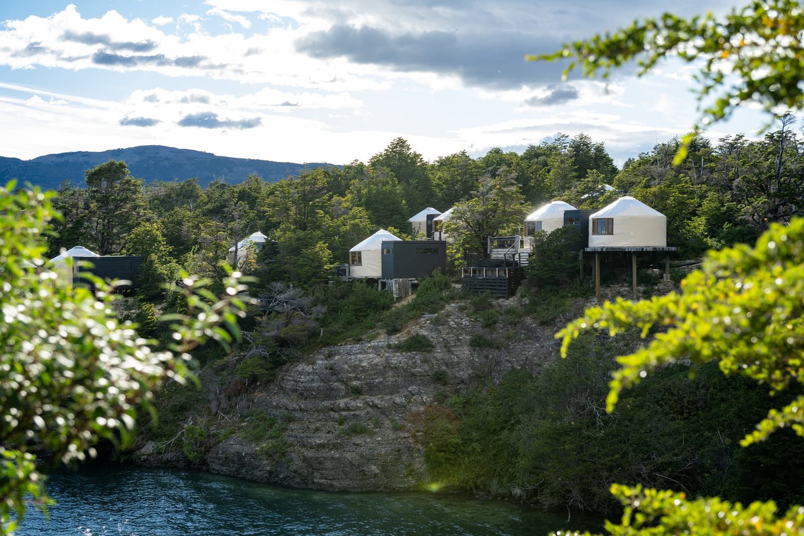 From the water, the camp looks like it grew there. It sort of did. Photo: Patagonia Camp Several white-roofed patagonia camp yurts perched on a rocky forested cliff above turquoise lake water, with dark mountains and a partly cloudy sky in the background
