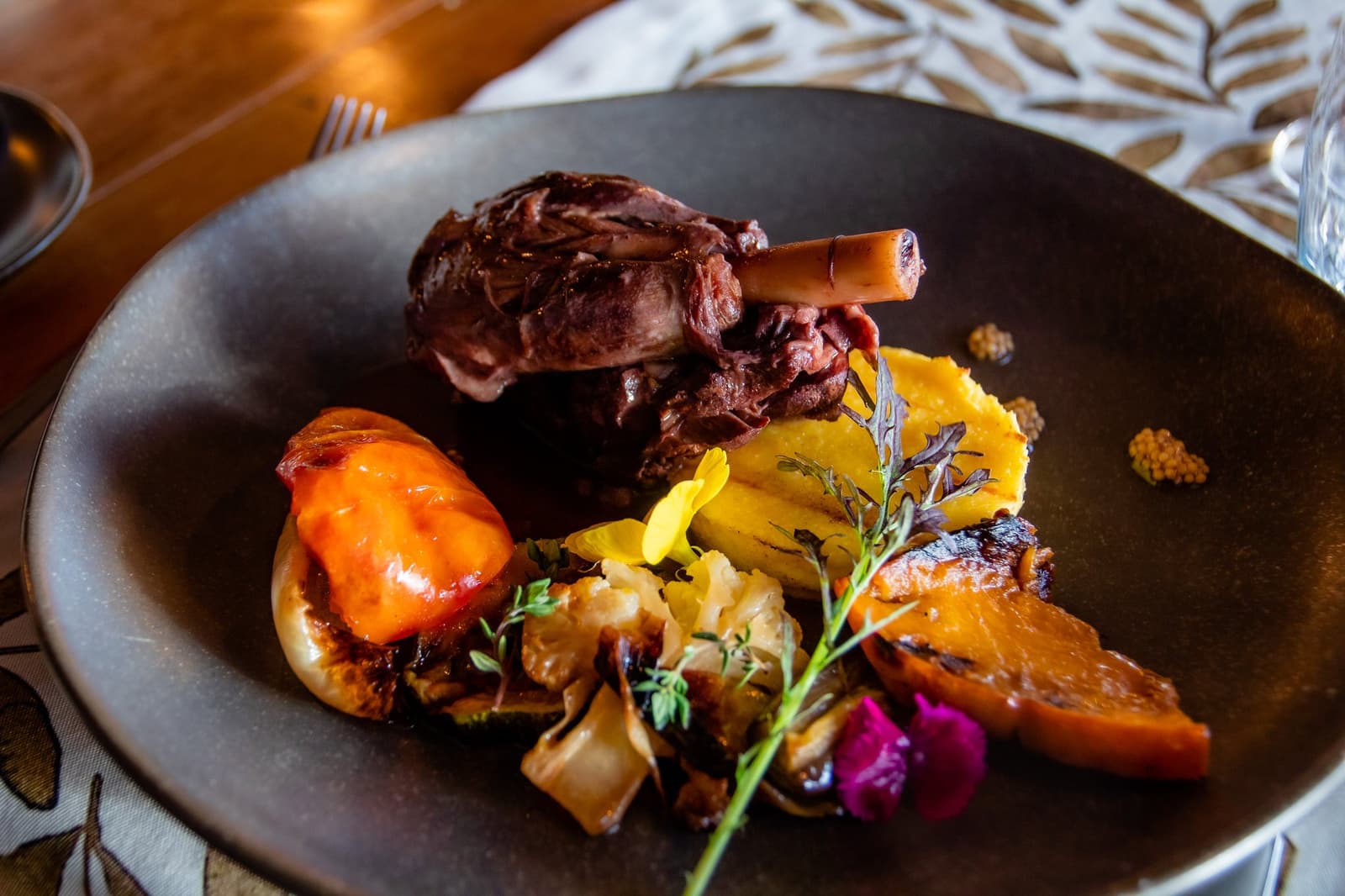 Ranch lamb, slow-cooked until the bone tells you it's ready. A braised lamb shank on a dark plate at Estancia Cerro Guido, served with golden potato purée, roasted tomato, caramelized citrus, charred cauliflower, fresh thyme, and edible flowers