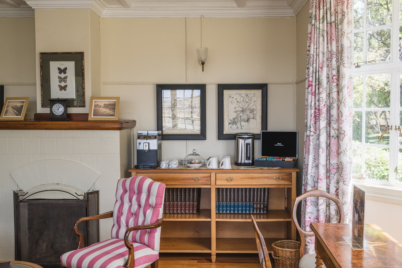 Books on the shelf, tea on the sideboard, and nowhere you need to be. Photo: CARLOS DIAZ Cozy sitting room at Estancia Cerro Guido with a wooden sideboard holding a coffee and tea station, framed botanical prints, a striped pink armchair beside a tiled fireplace, and tall windows with floral curtains overlooking the garden