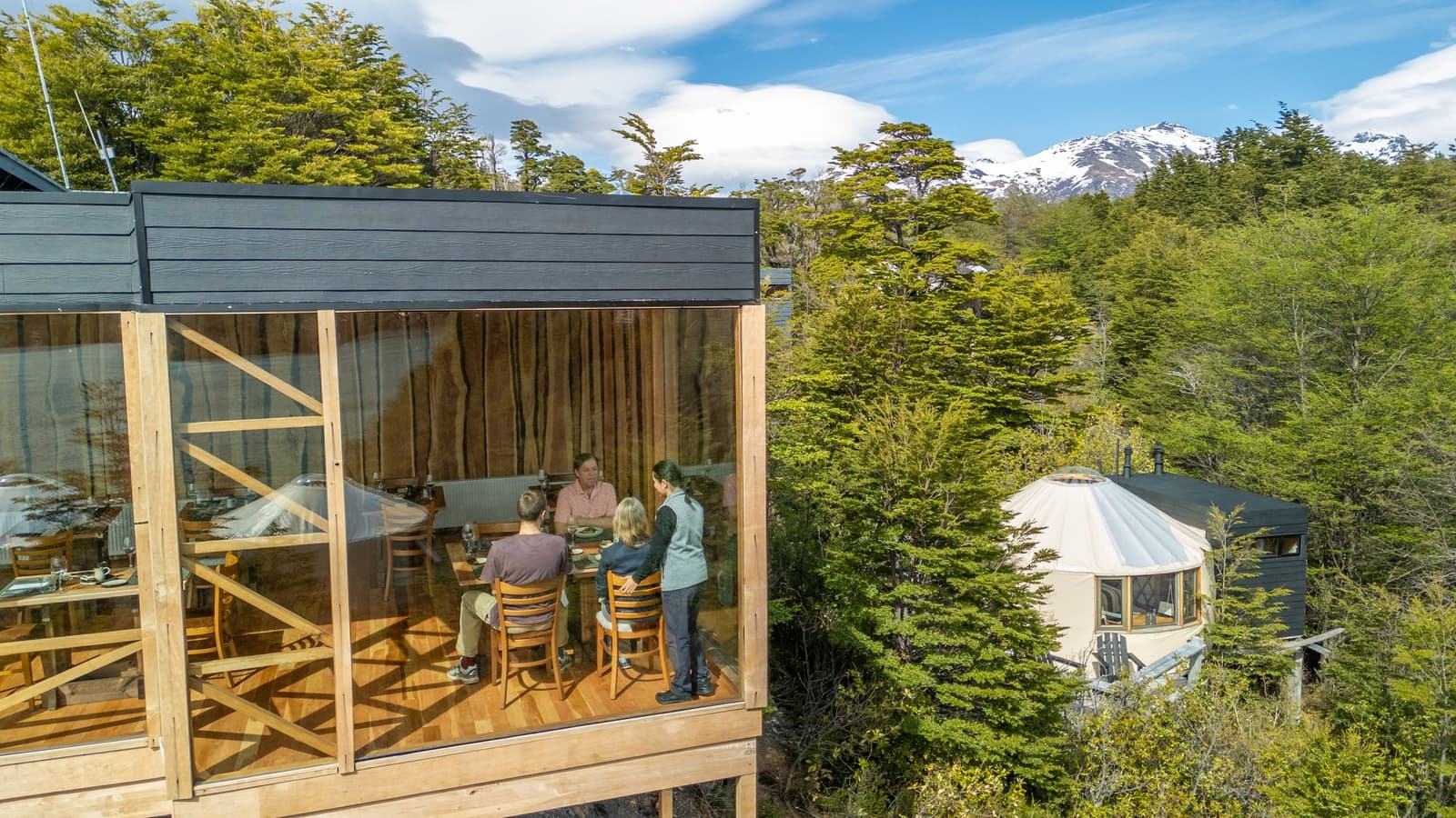 Good food, good company, and snow-capped peaks just outside the glass. Photo: Patagonia Camp Aerial view of the Patagonia Camp glass-walled dining room on stilts, where guests share a meal around a wooden table while a staff member attends them, with a circular yurt, native lenga beech forest, and a snow-capped mountain visible in the background