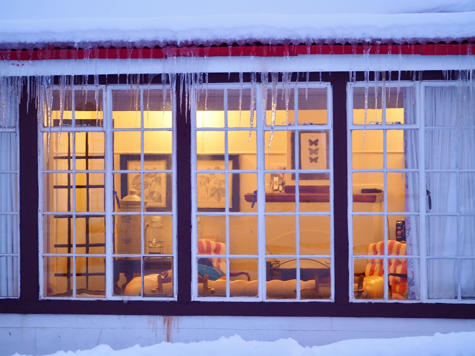 Cold outside, very much not cold inside. Warm golden light glows through large multi-pane windows of the estancia cerro guido lodge exterior at dusk, with icicles hanging from a snow-covered red roof and snow on the ground