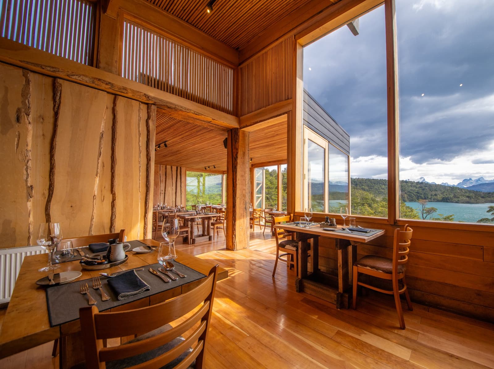 Every seat in the house comes with mountains, a lake, and no reservations required. Photo: Patagonia Camp Warm timber interior of the Patagonia Camp restaurant with set tables, wine glasses, and floor-to-ceiling windows framing a turquoise lake and distant mountain peaks under a dramatic cloudy sky