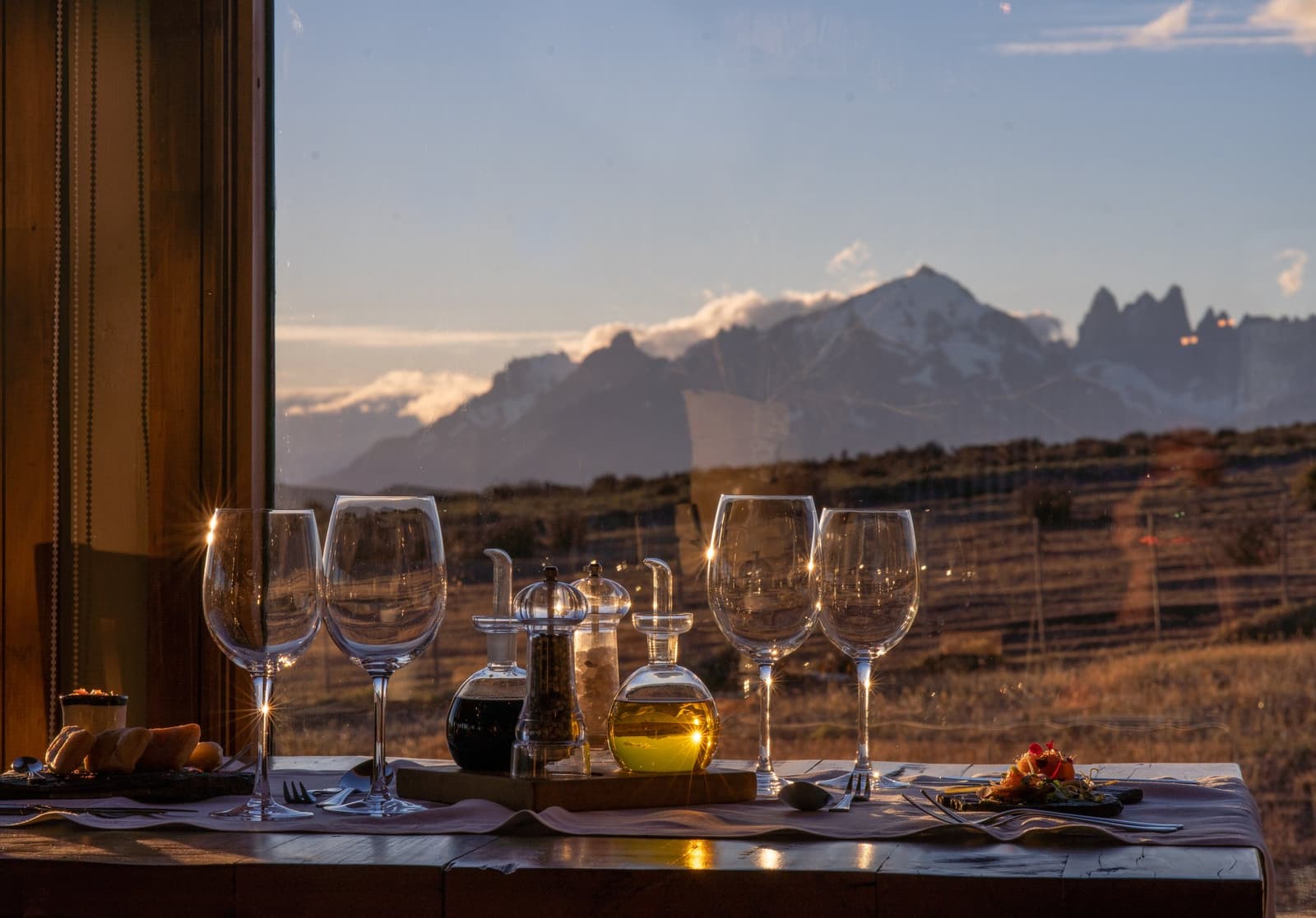 Dinner reservations: one table, four glasses, and those peaks doing all the talking. A set dining table with crystal wine glasses, olive oil, and bread at Estancia Cerro Guido, with a panoramic window framing the jagged peaks of the Paine Massif across the golden steppe at dusk