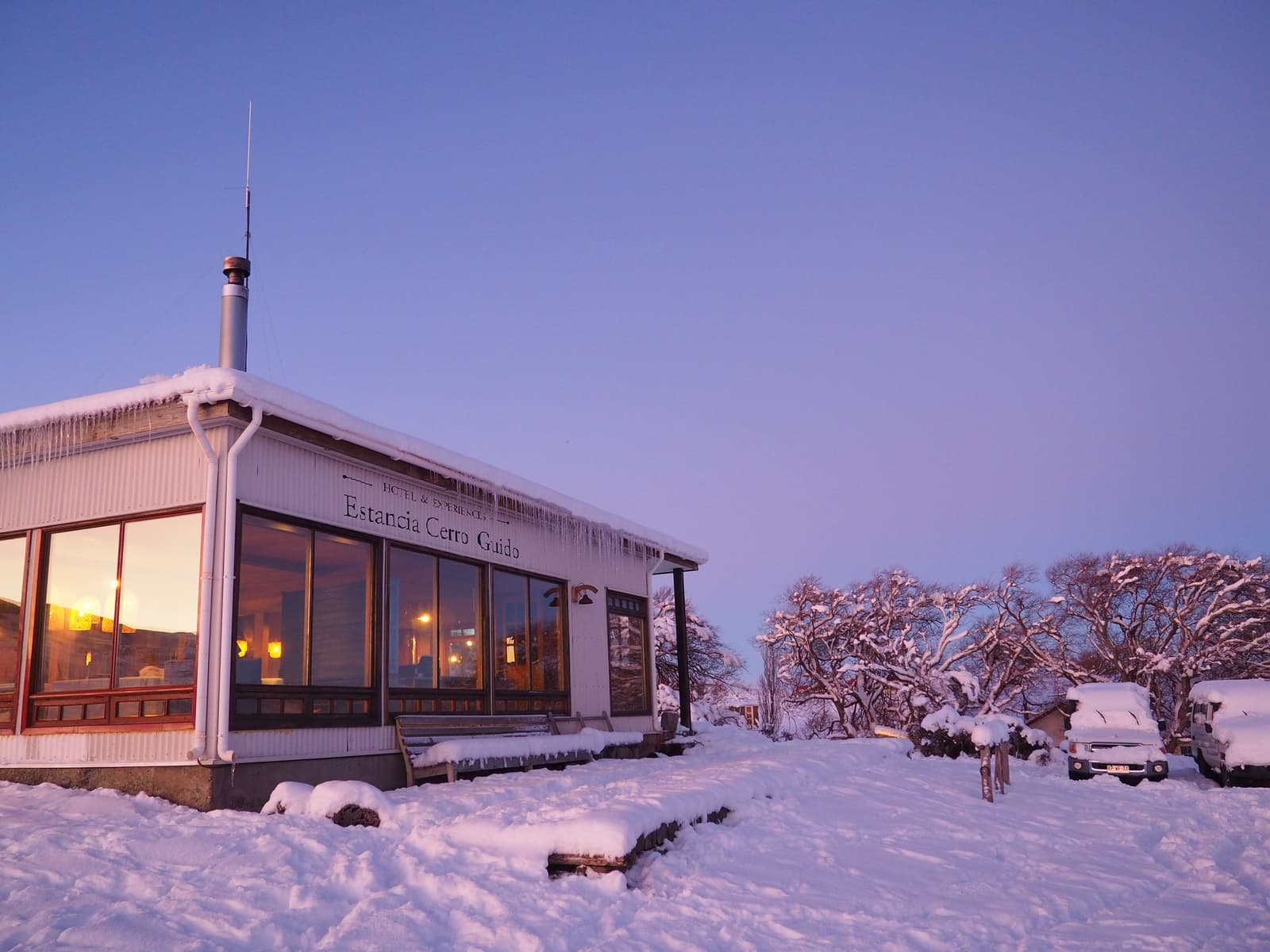 Icicles on the eaves, lights on inside. The estancia earns its keep on a cold night. Photo: Estancia Cerro Guido hotel and experience building at dusk in winter, with icicles hanging from a snow-covered roof, warm interior lights glowing through large windows, and snow-laden trees visible in the purple evening sky