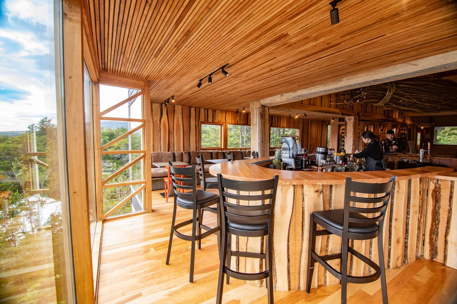 Post-hike drinks hit differently when the forest is right there through the glass. Photo: Patagonia Camp Patagonia Camp bar and lounge interior with a curved live-edge wooden counter, bar stools, floor-to-ceiling windows overlooking native forest and a cloudy sky, and two staff members working behind the bar