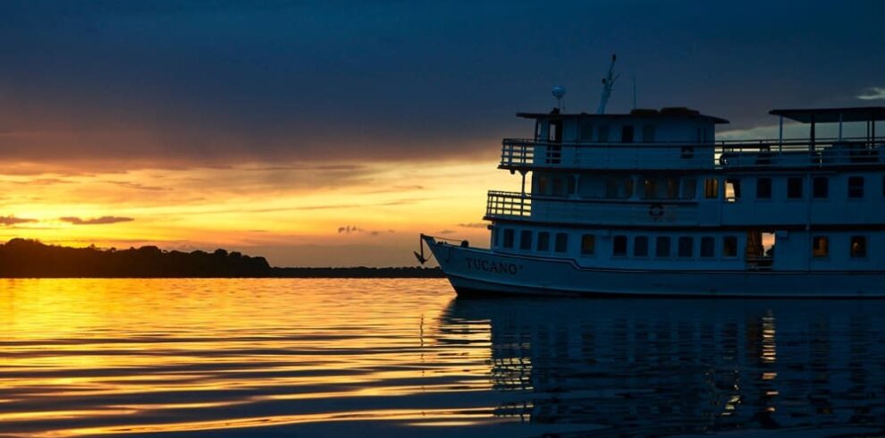 The silhouette of the Tucano Amazon river cruise boat sailing during a vibrant sunset over the water.