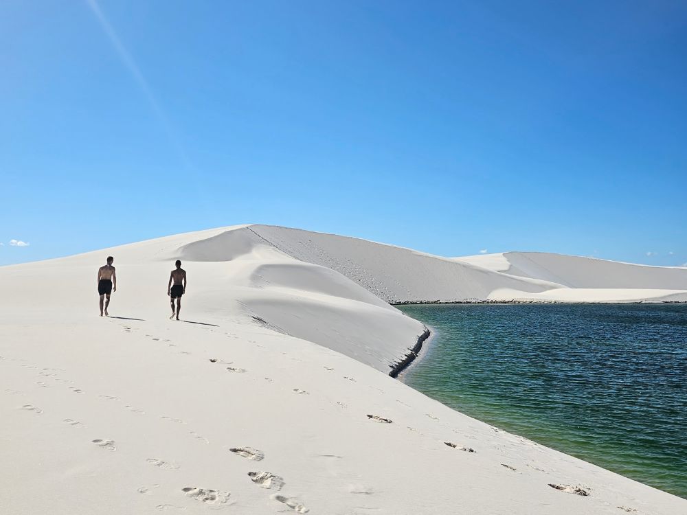 Zwei Personen spazieren am Ufer einer leuchtend blauen Lagune, umgeben von den weißen Dünen der Lençóis Maranhenses.