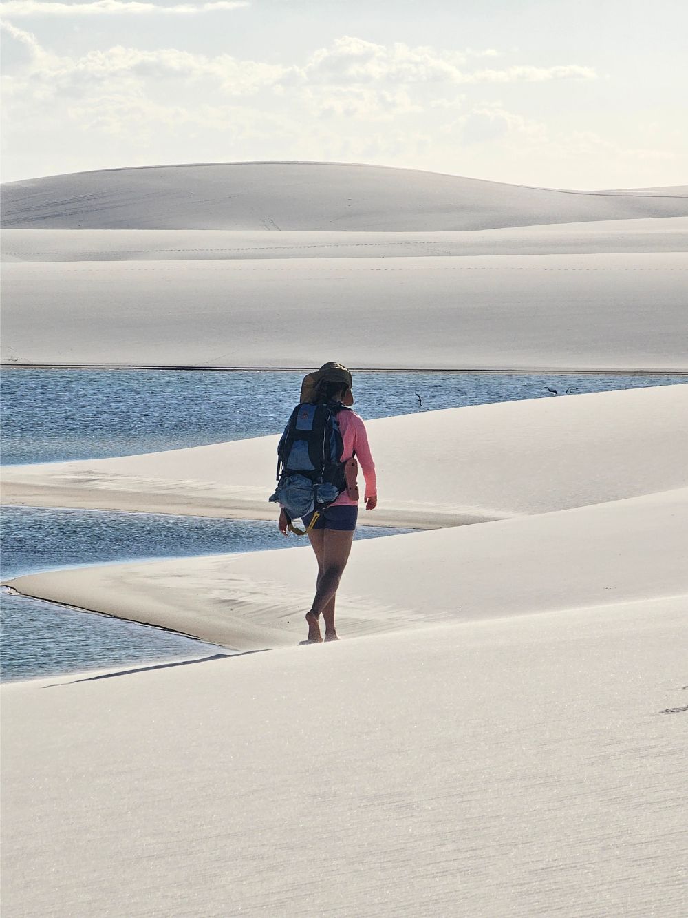 Ein Rucksacktourist wandert barfuß über die weißen Sanddünen in der Nähe einer blauen Regenwasserlagune im Lençóis Maranhenses Nationalpark.