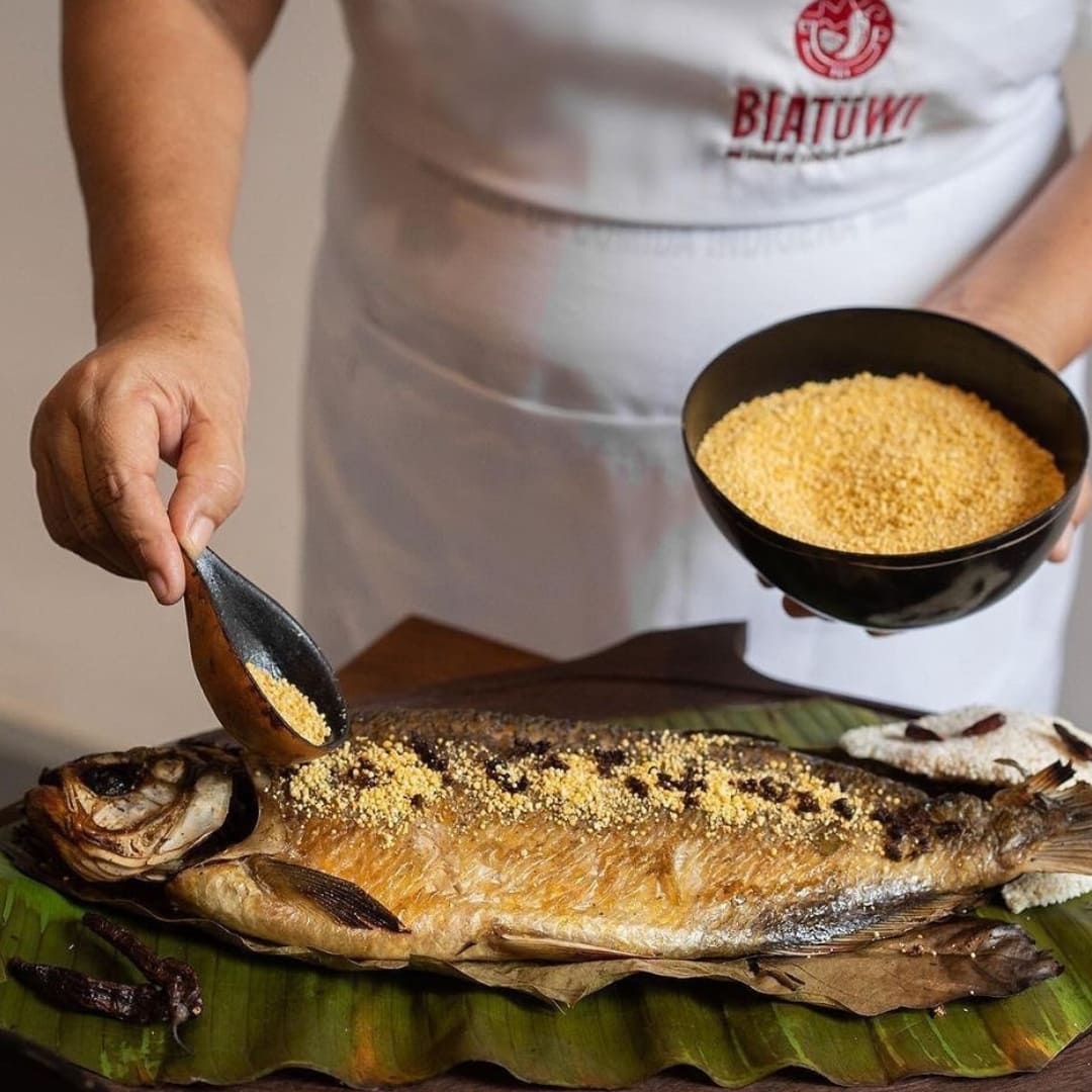 Close-up van een persoon die maniokmeel strooit over een geroosterd visgerecht geserveerd op een bananenblad.