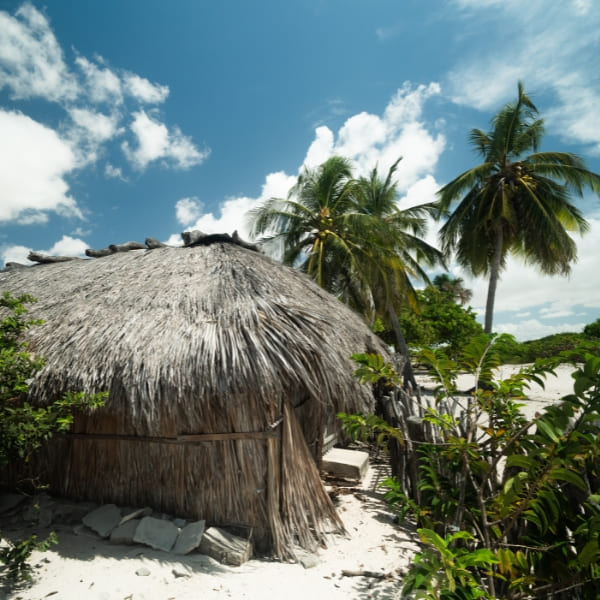 Eine Person ruht in einer orangefarbenen Hängematte unter einem rustikalen, offenen Strohdach-Unterstand, der mit bunten Hängematten gefüllt ist.