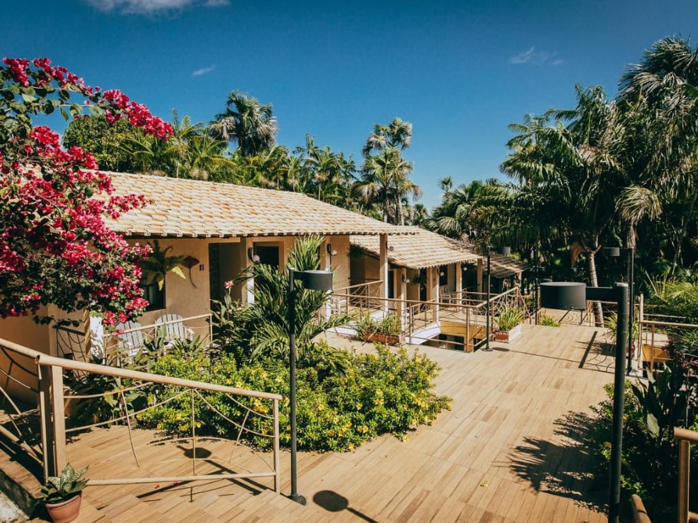 Sonniger Außenbereich einer rustikalen Pension mit blühendem Garten, Holzrampen und Treppen, die zu den Zimmerbalkonen führen.