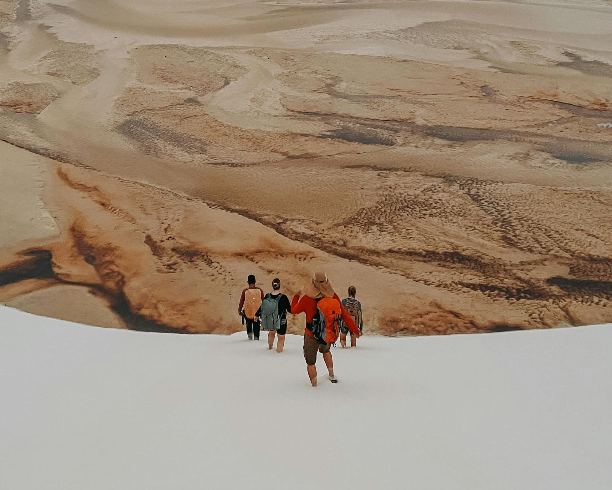 Un groupe de touristes traverse à pied le lit asséché d'une lagune brune, entourée de grandes dunes de sable blanc, en pleine saison sèche.